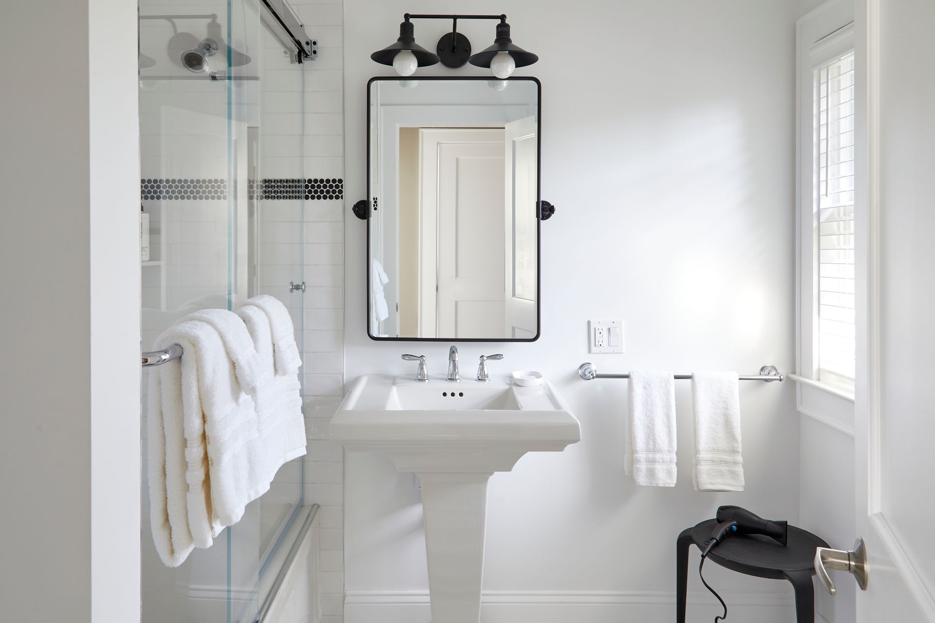 Bright white bathroom with pedestal sink, mirror, glass shower, and towels hanging nearby.