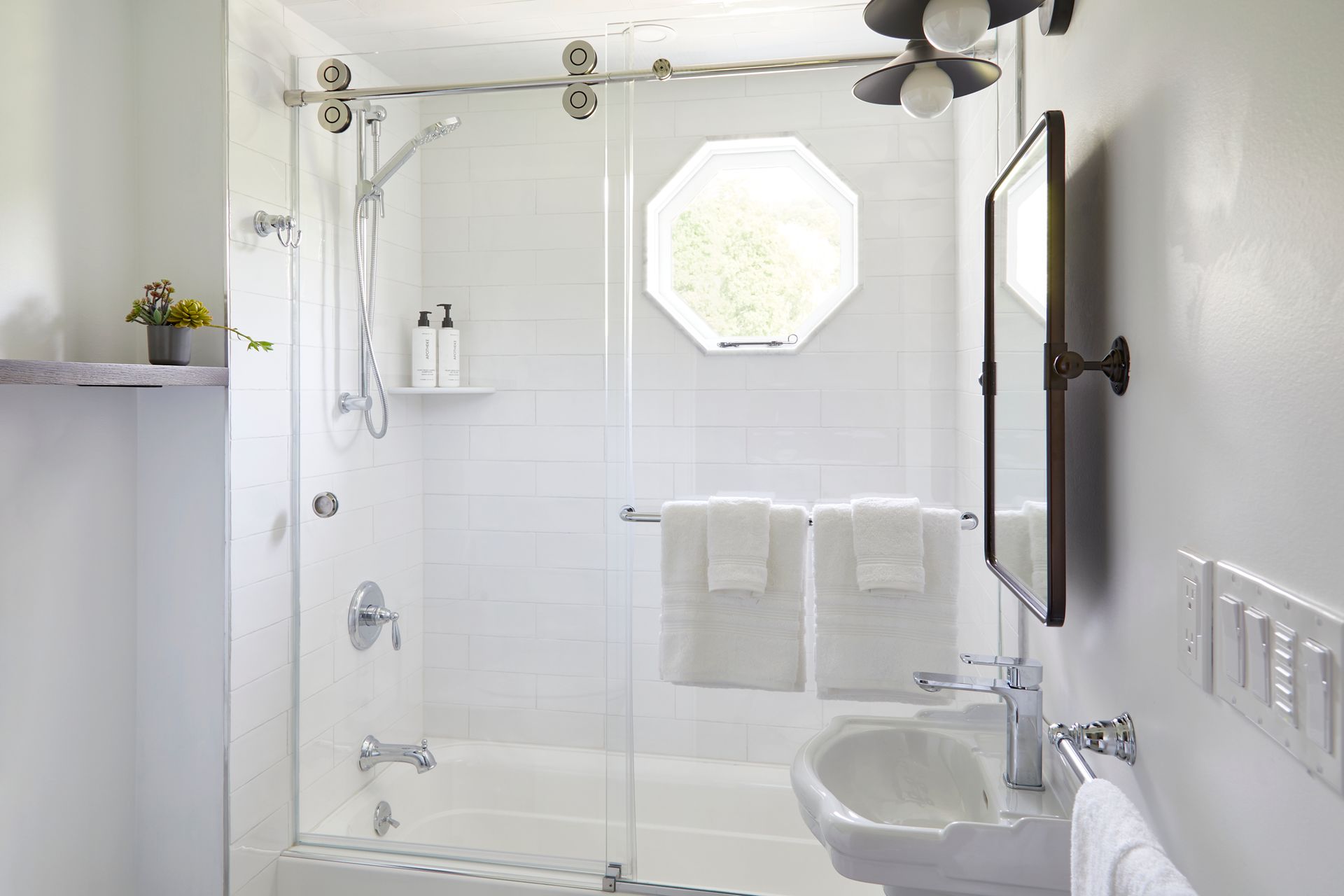 Bright white bathroom with glass shower, towel bars, sink, and octagonal window.