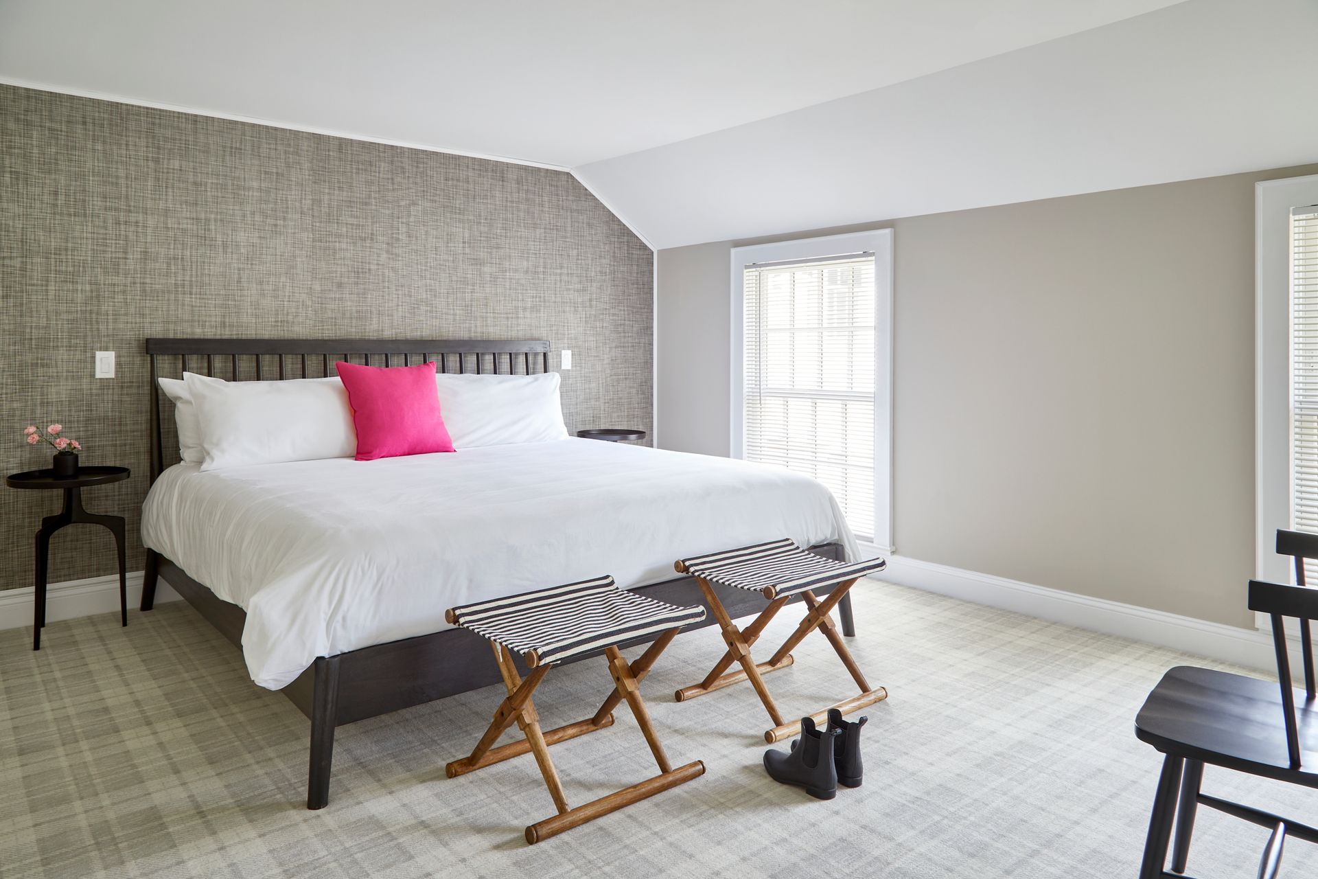 Bright bedroom with a large bed, pink pillow, wicker stools, and windows with white blinds