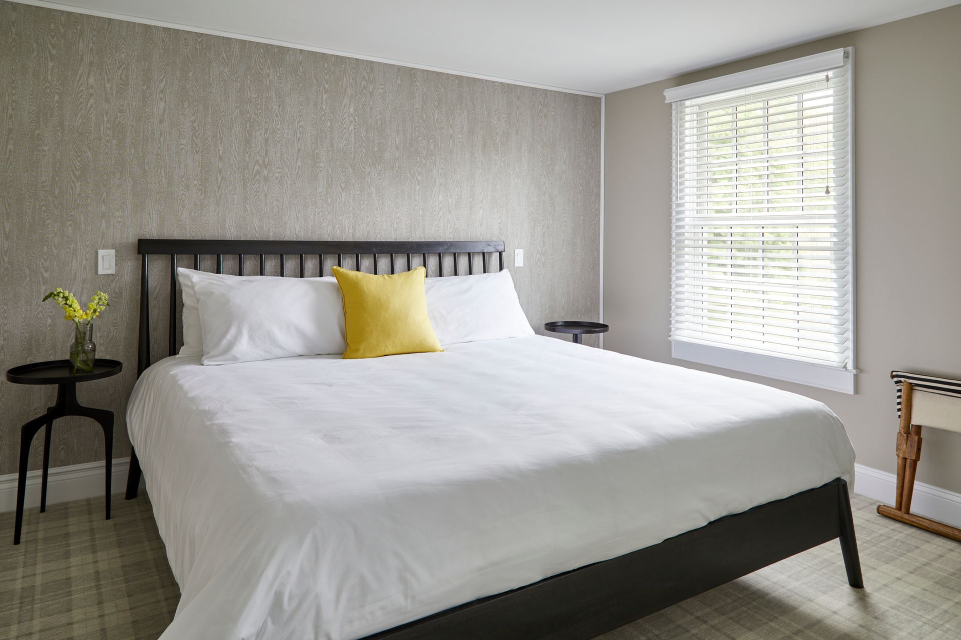 Bright bedroom with a made bed, white bedding, yellow accent pillow, and a window with blinds