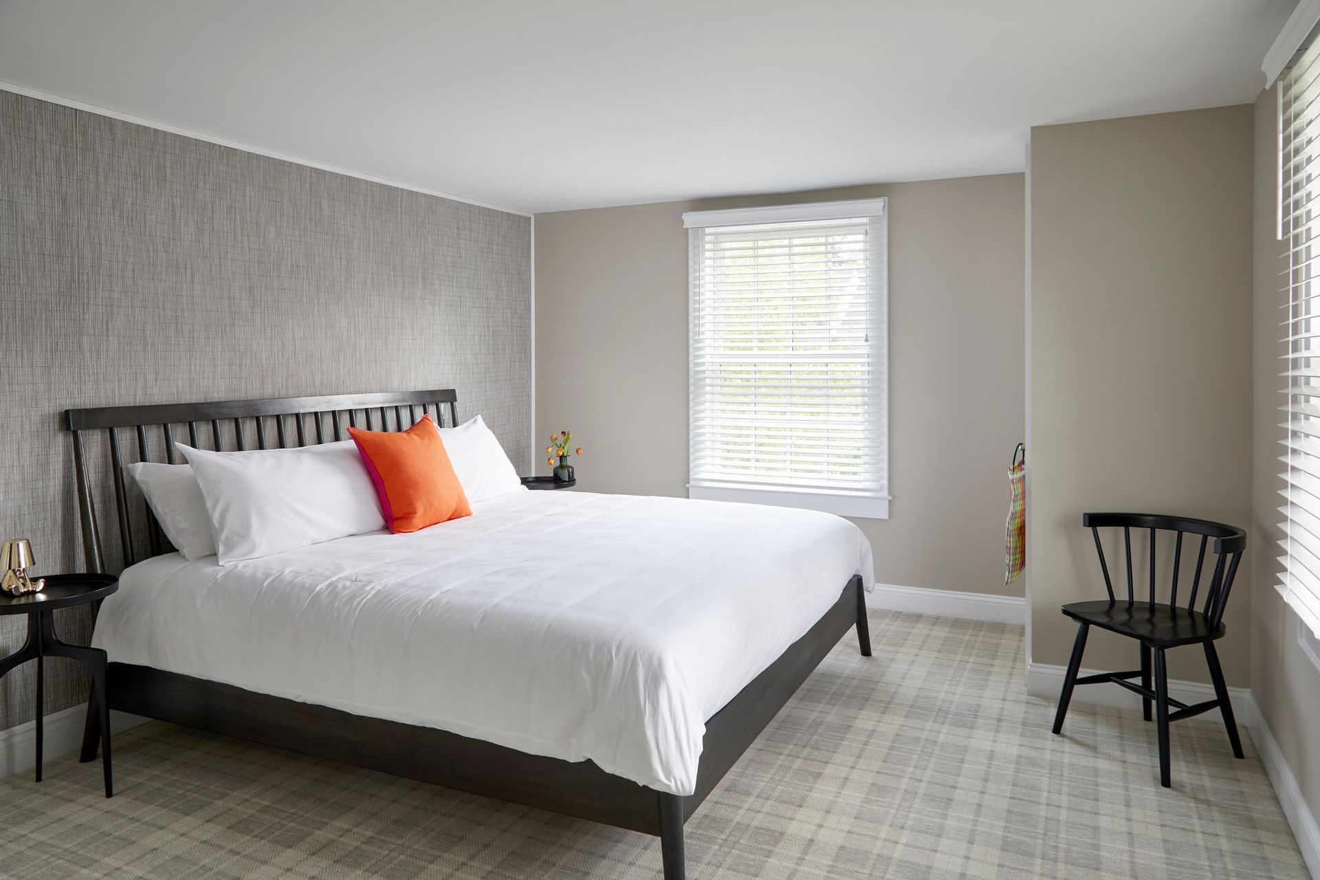 Bright bedroom with a made bed, white bedding, orange pillow, gray walls, and two black chairs