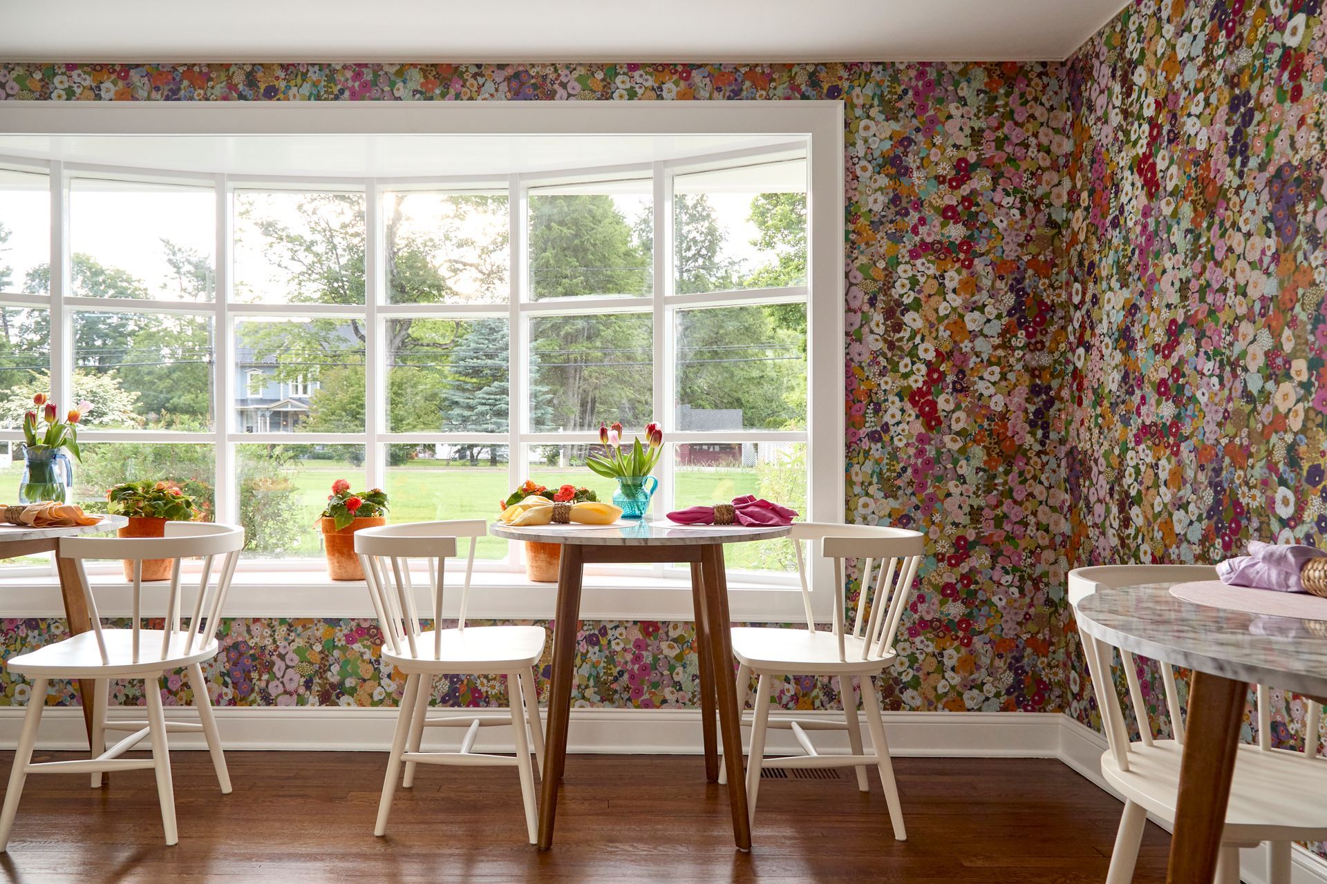 Bright dining room with floral wallpaper, large windows, white chairs, and a wooden table by the window.