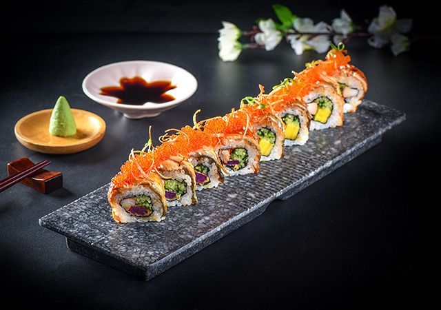 Sushi rolls topped with orange garnish on a black platter, with soy sauce and wasabi on a dark table