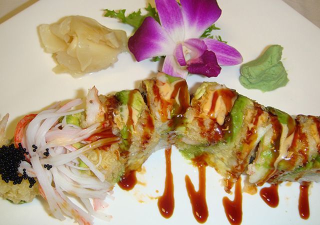 Sushi roll on a white plate, topped with sauce, with shredded garnish and purple orchid decoration