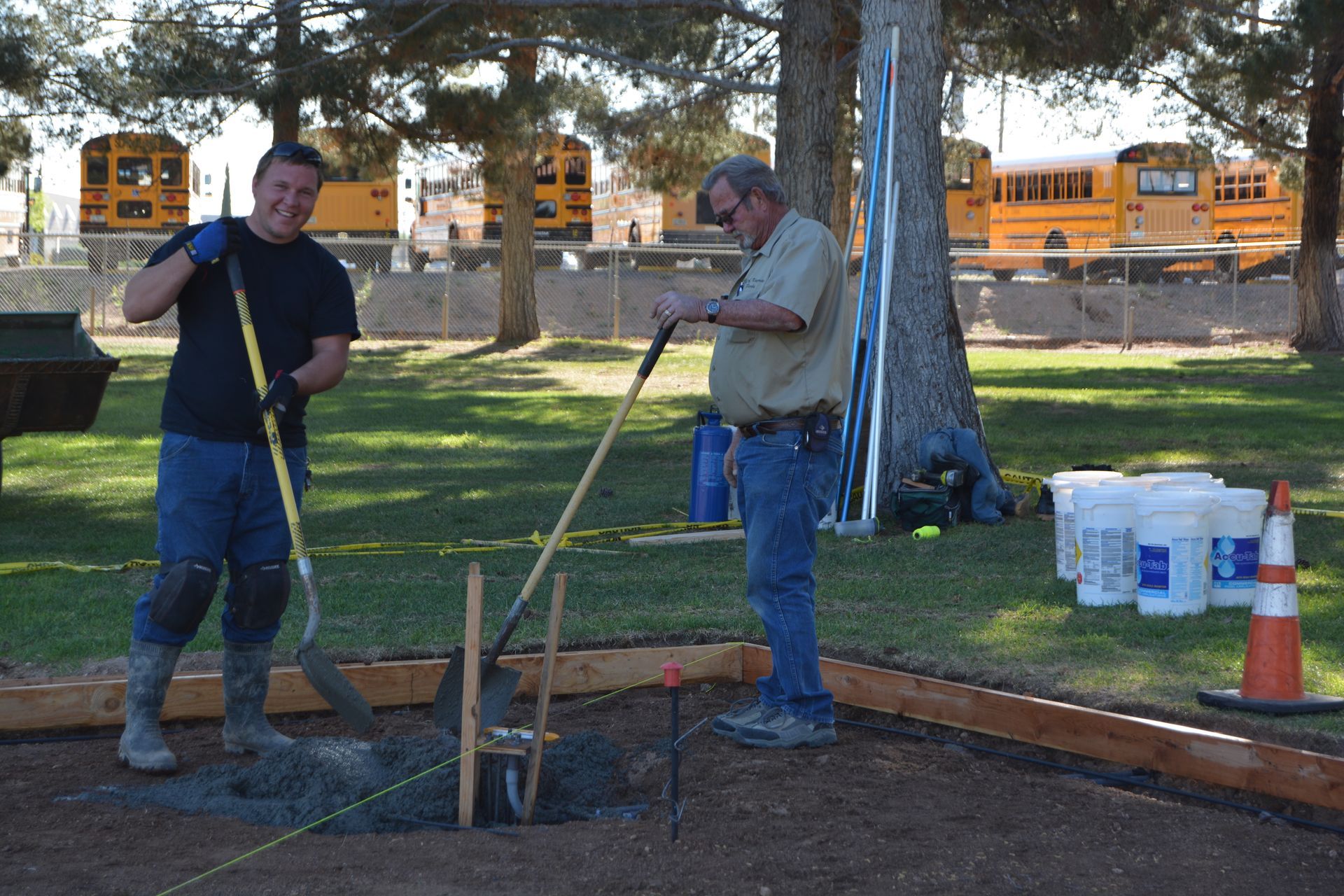 Two men are working in a field with school buses in the background