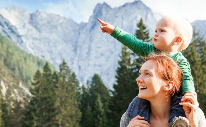 A woman is carrying a baby on her shoulders in front of a mountain.