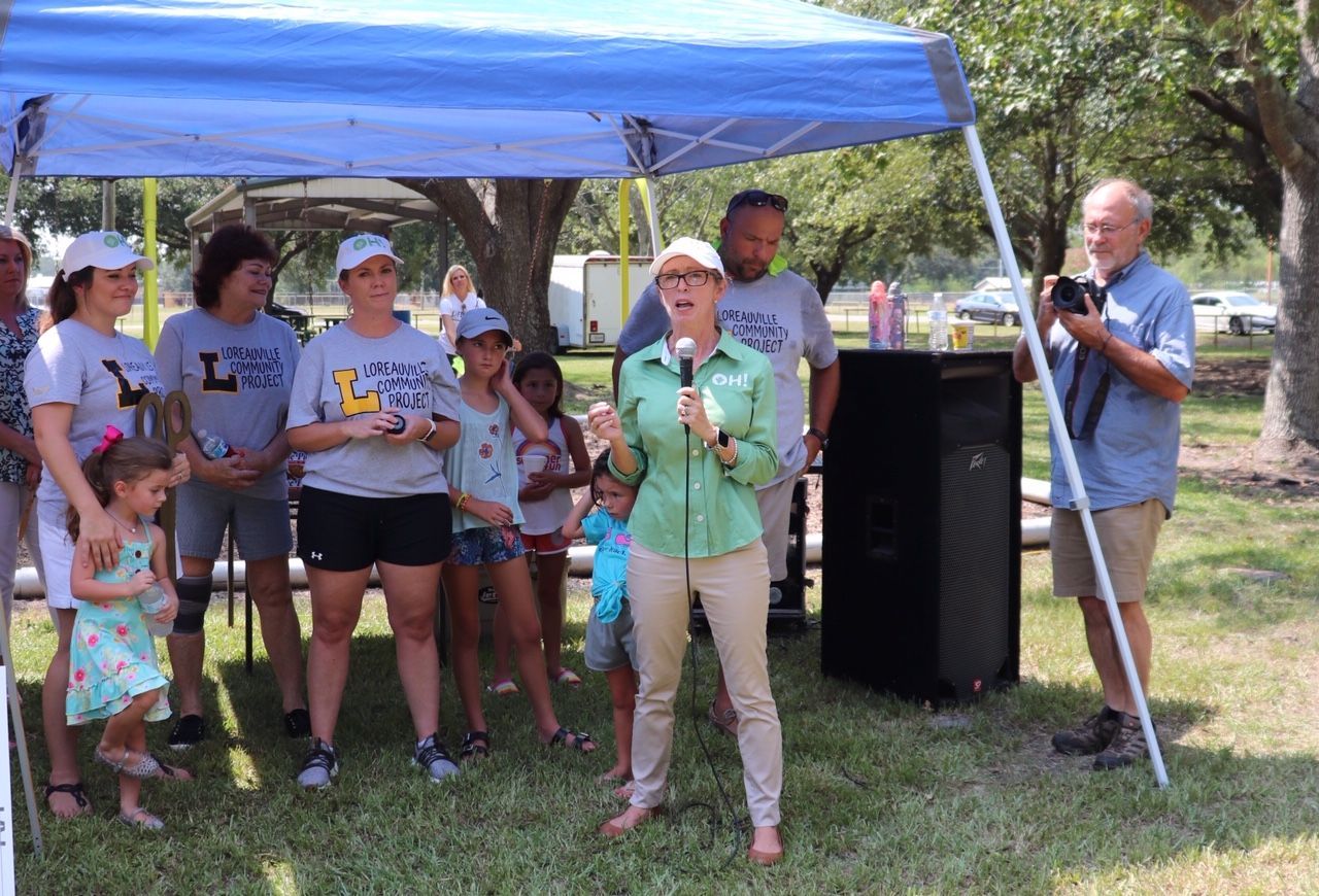 A woman is speaking into a microphone in front of a group of people under a blue tent.