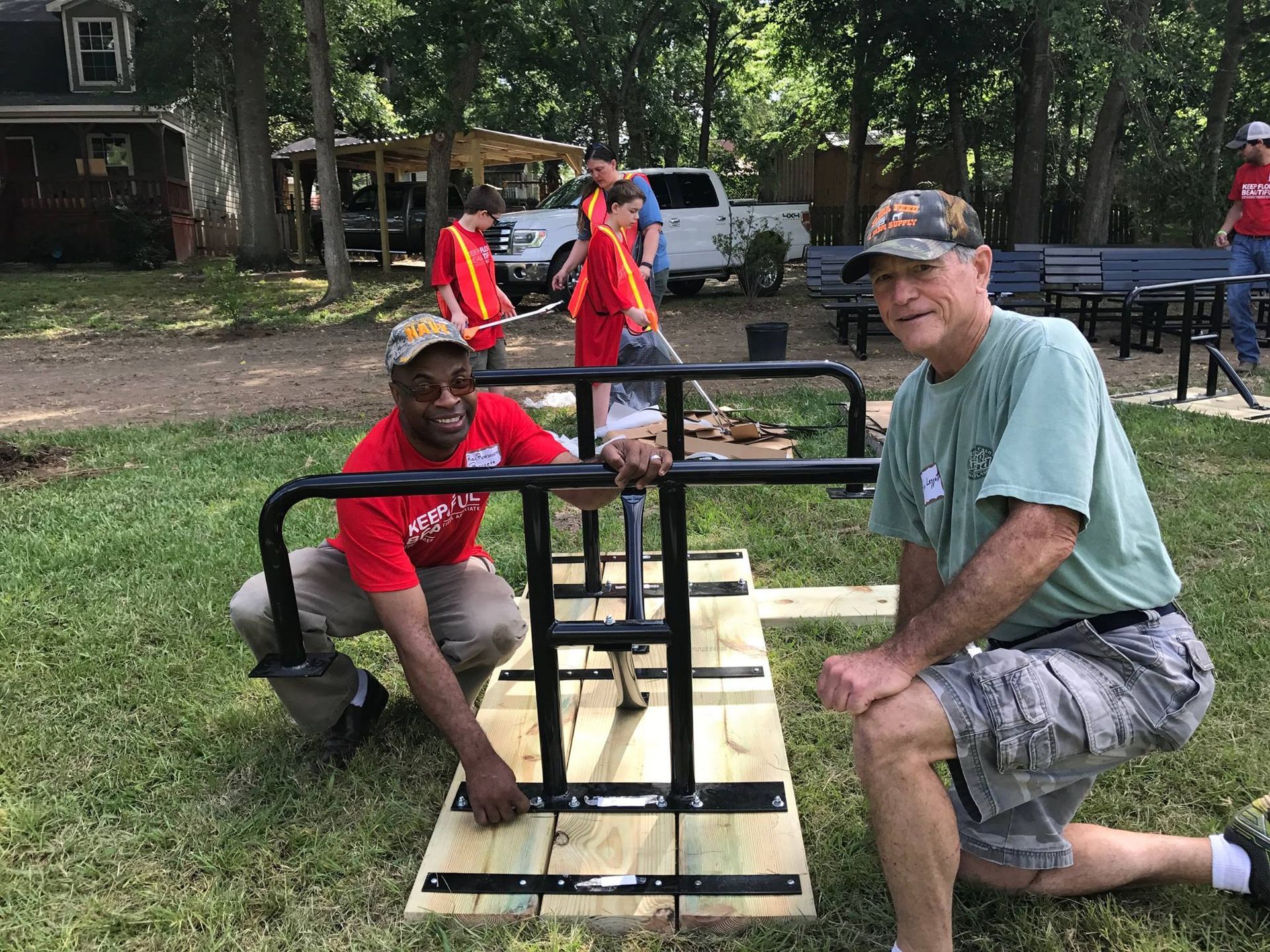 Two men are working on a wooden table in a park.