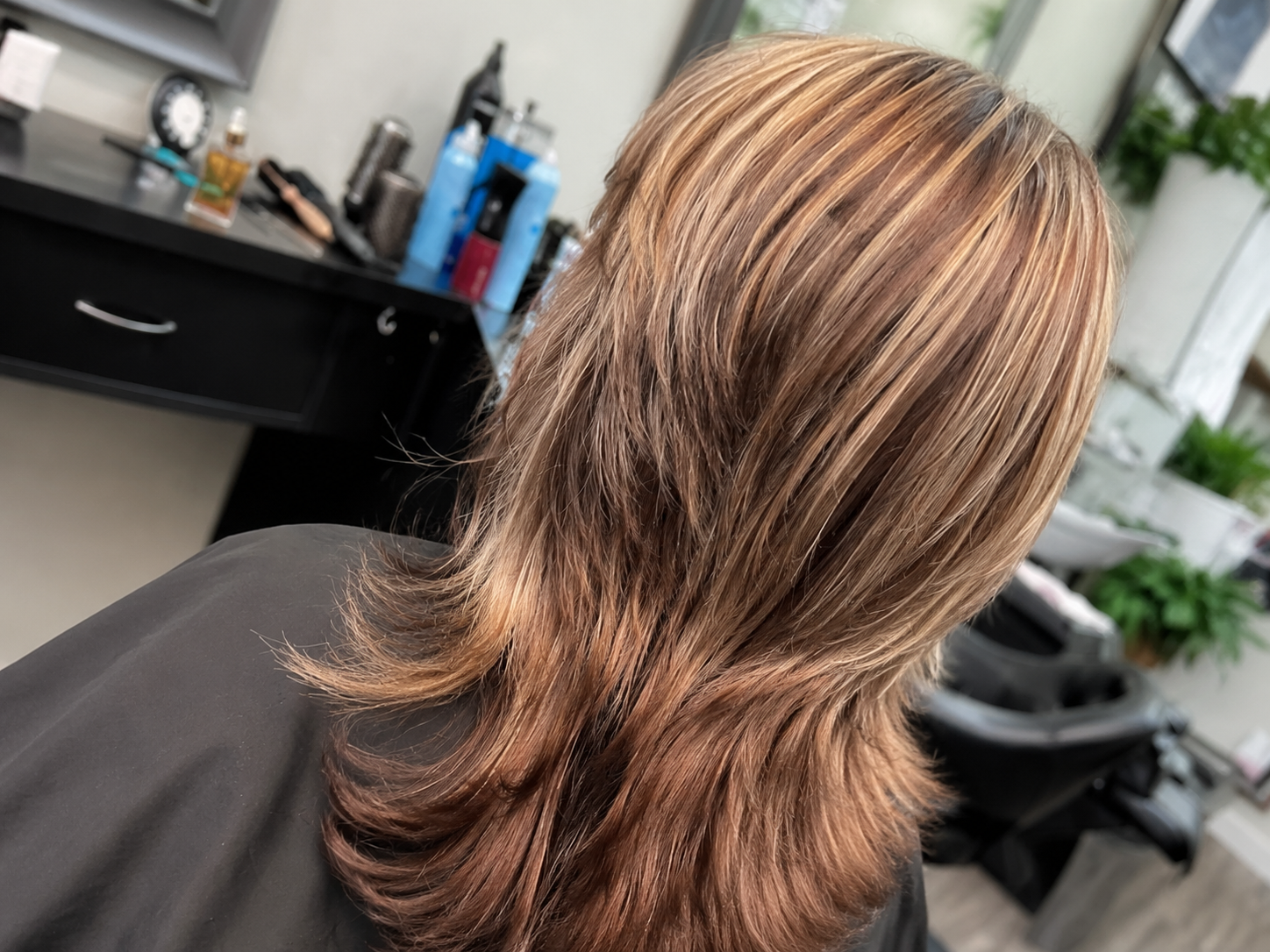 Woman with wavy, highlighted hair in a salon chair, hair cape on.