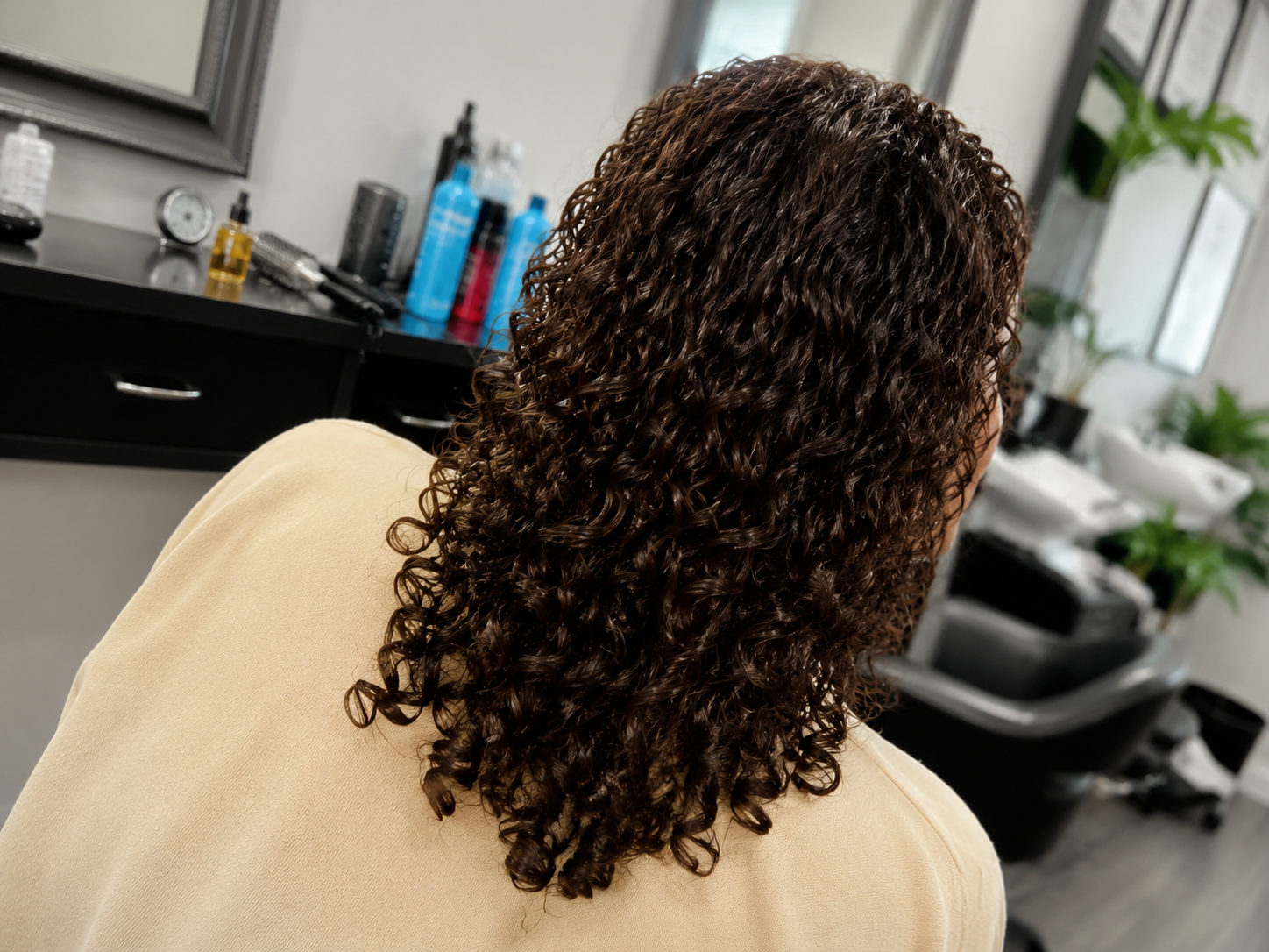 Person with curly brown hair sitting in a salon chair, viewed from behind.