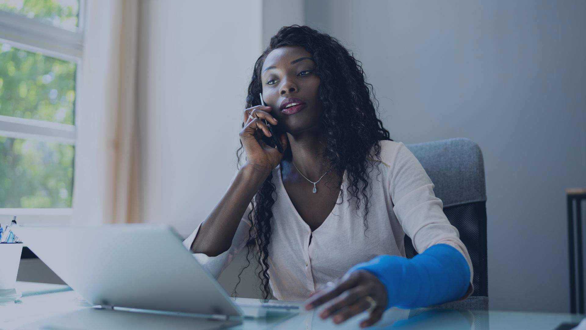 a woman with a cast on her arm is sitting at a desk talking on a cell phone talking about Worker's Compensation.