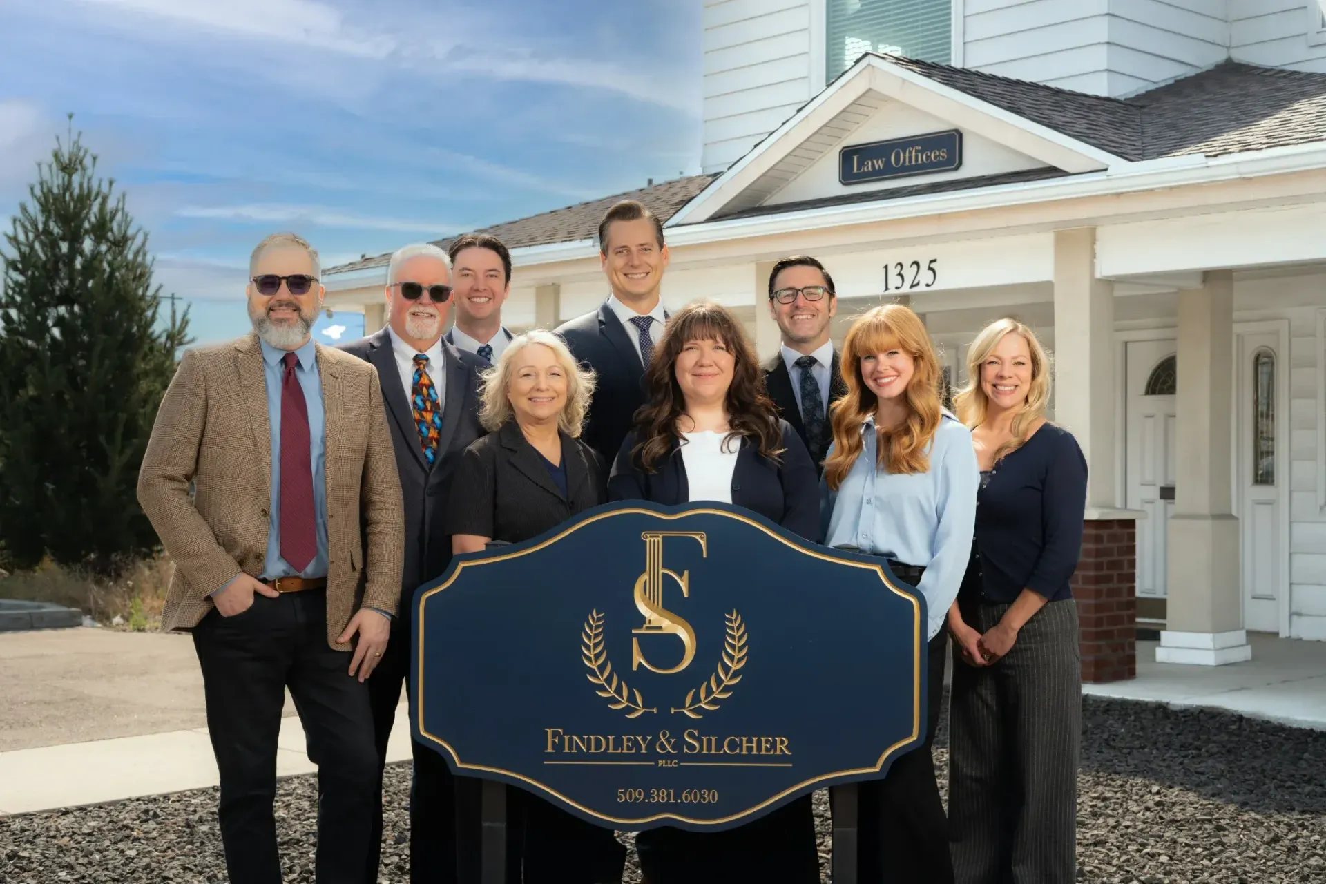 Findley & Silcher Law team posing outside the law firm behind their blue and gold business logo sign