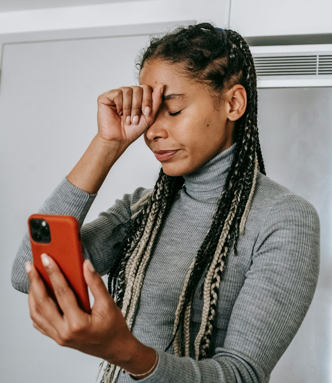 Woman in gray sweater holding a red phone and rubbing her eye while looking at the screen