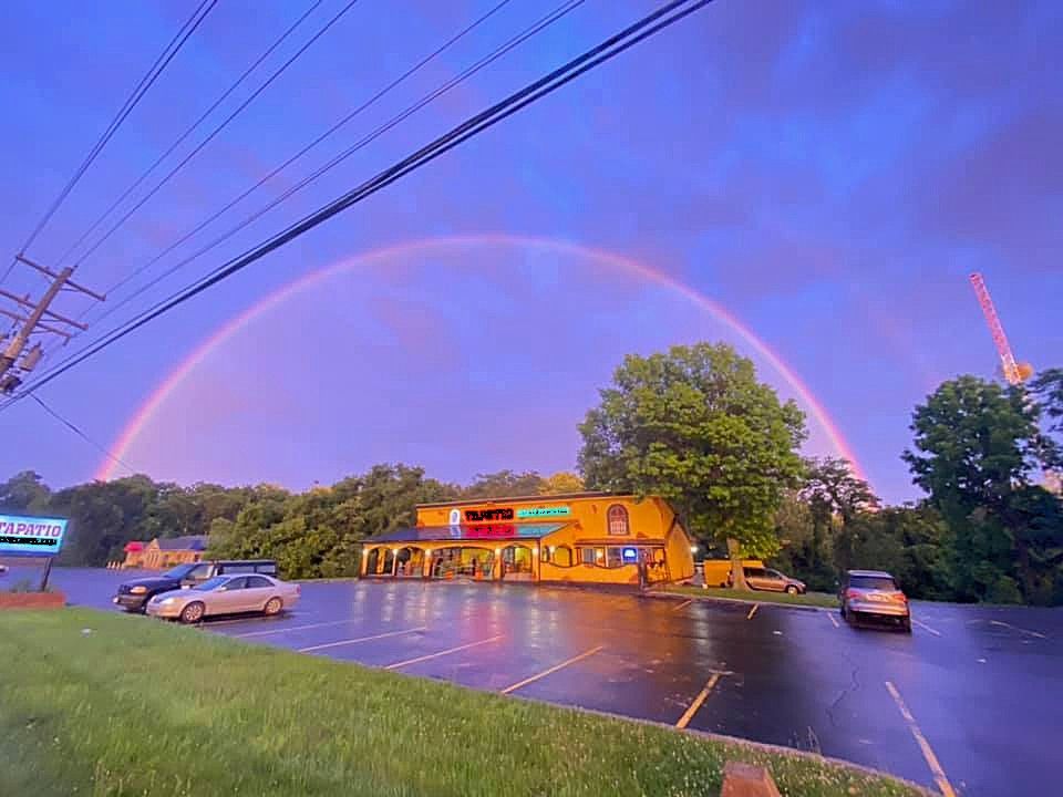 Rainbow arcs over a yellow building with a parking lot; cars parked. Trees and power lines in view.
