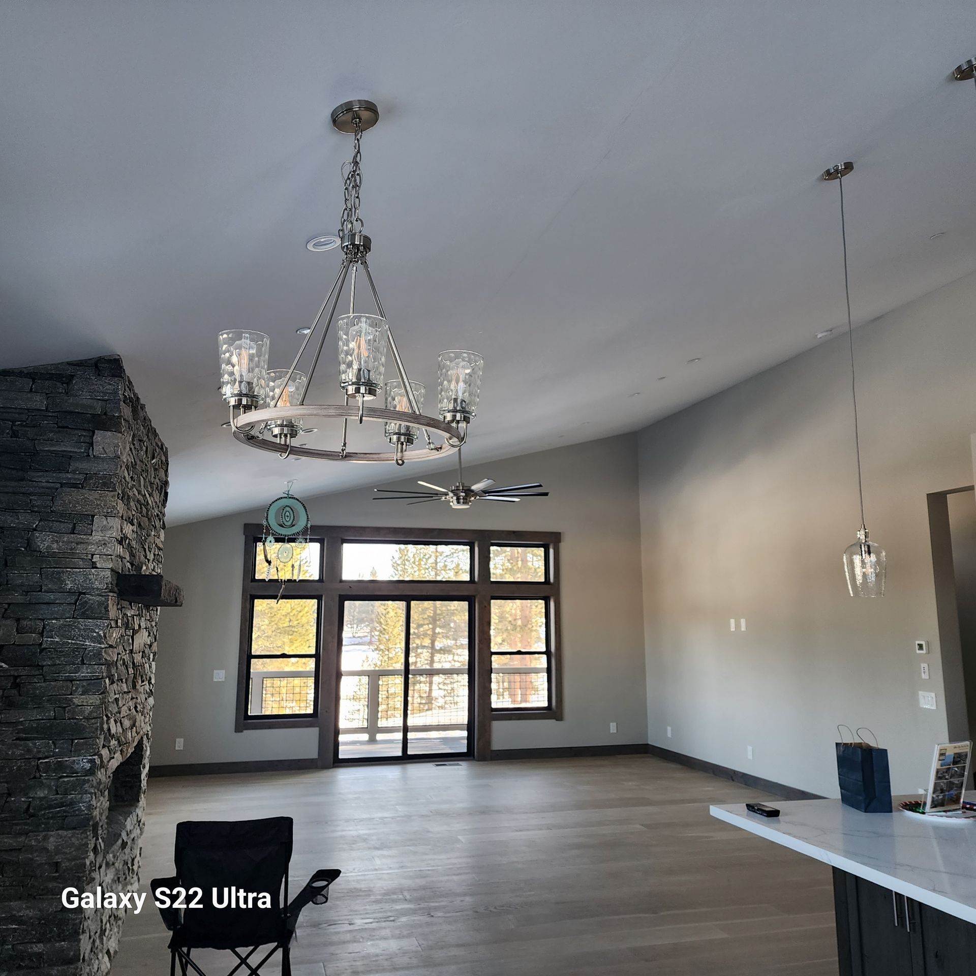 Empty modern living room with stone fireplace, chandelier, large windows, and open front doors.