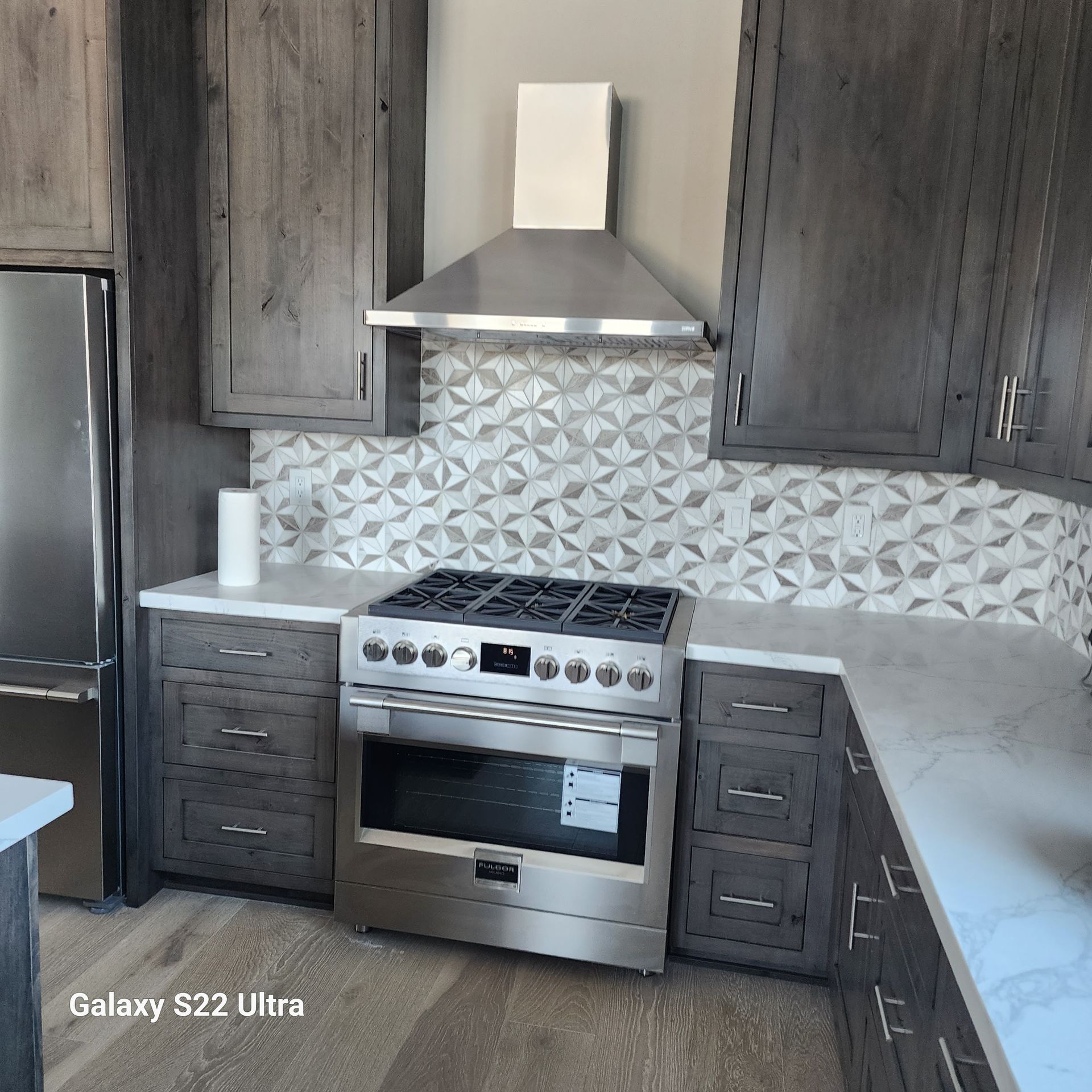 Modern kitchen with stainless steel stove, hood, and patterned backsplash between dark cabinets and white countertops
