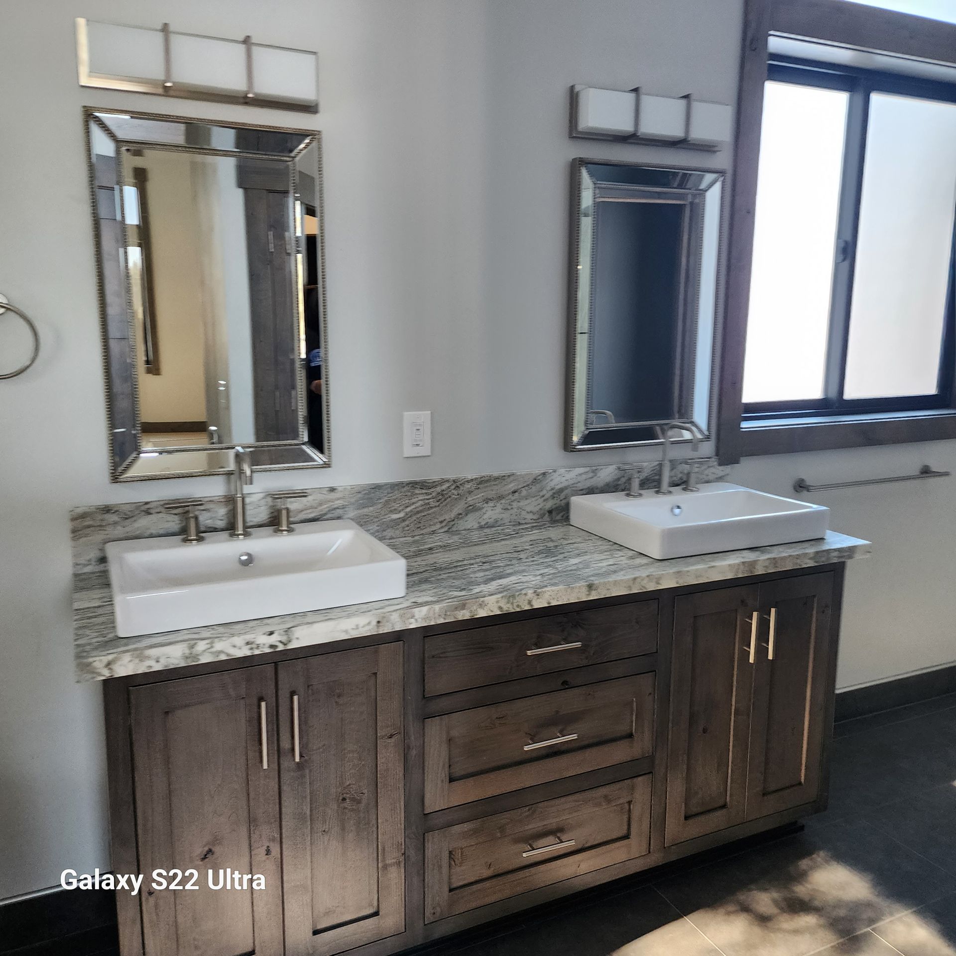 Bathroom vanity with double sinks, granite countertop, and two framed mirrors beside a window.
