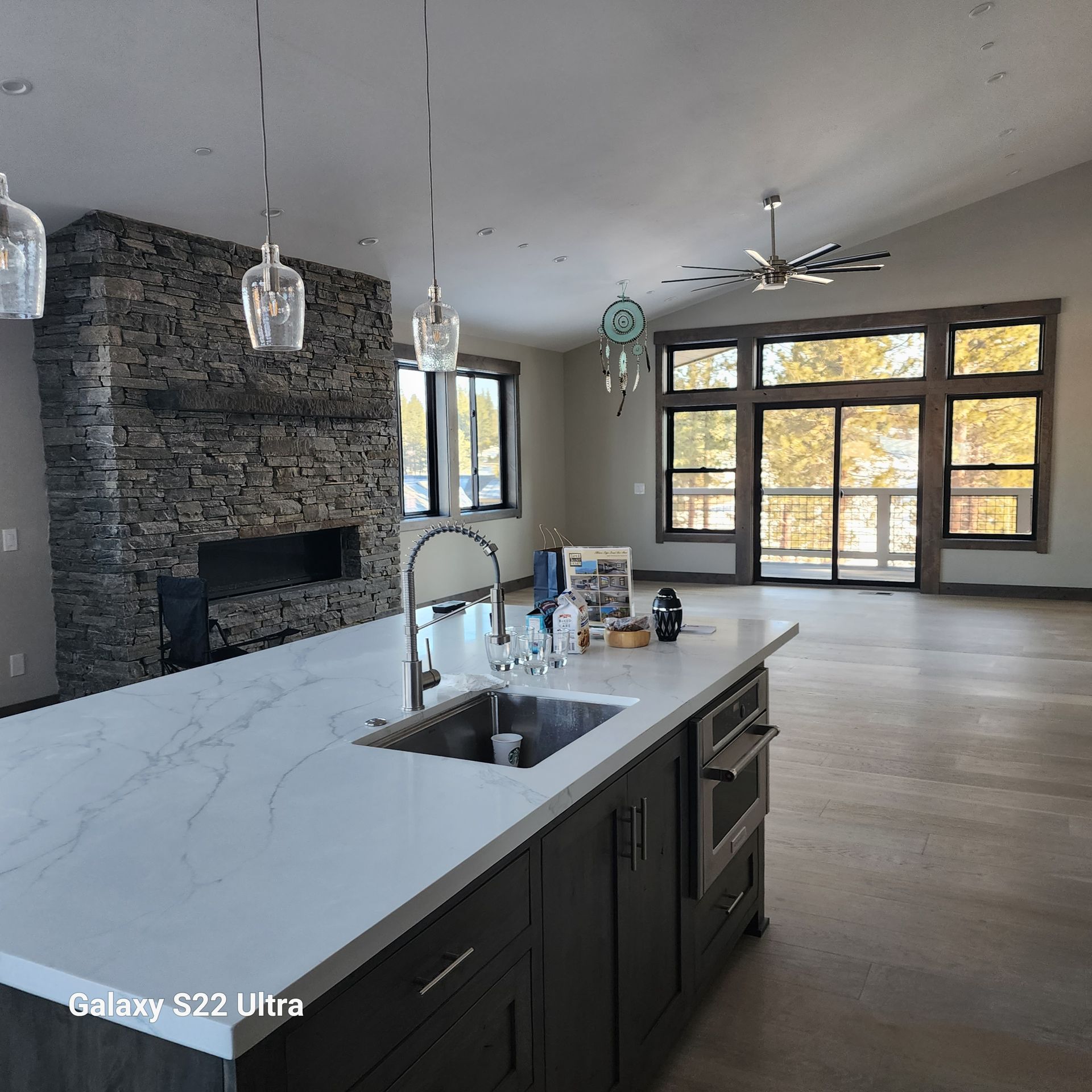 Modern open-concept kitchen with dark cabinets, white island, stone fireplace, and large windows overlooking autumn trees