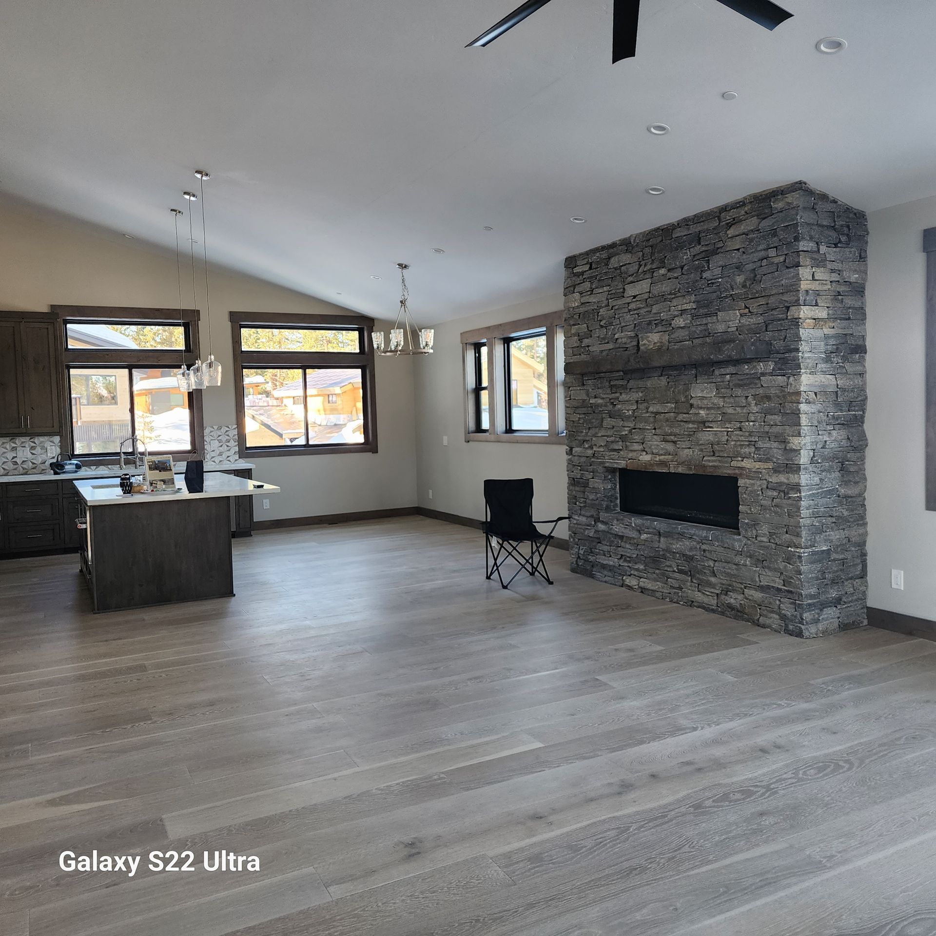 Open-concept living room with stone fireplace, kitchen island, large windows, and light wood floors.