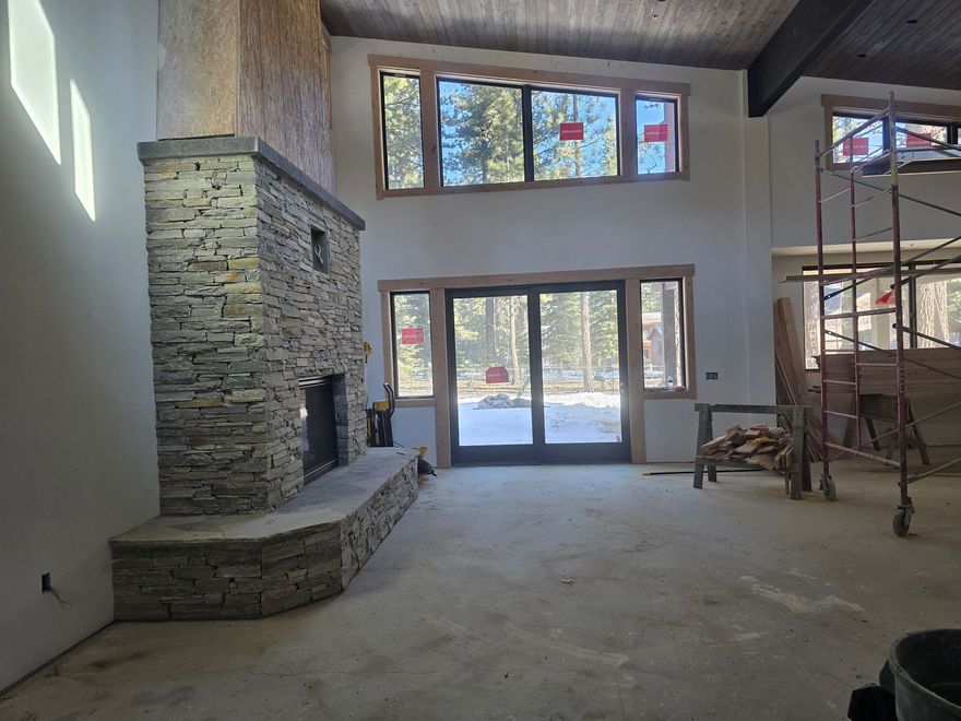 Empty modern living room with stone fireplace, tall windows, and sliding glass doors.
