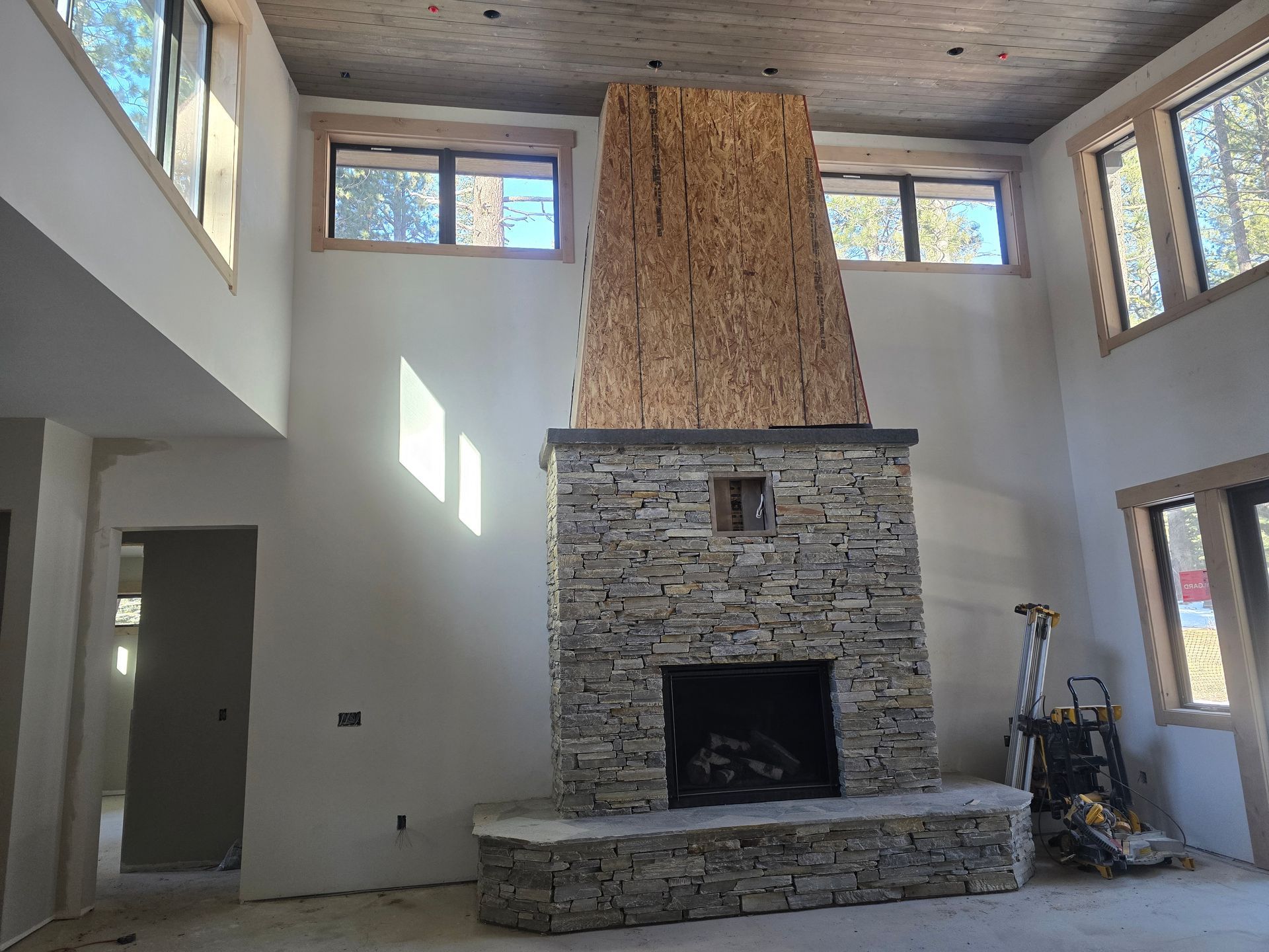 Interior with a tall stone fireplace, high windows, and a ladder in a bright room under construction