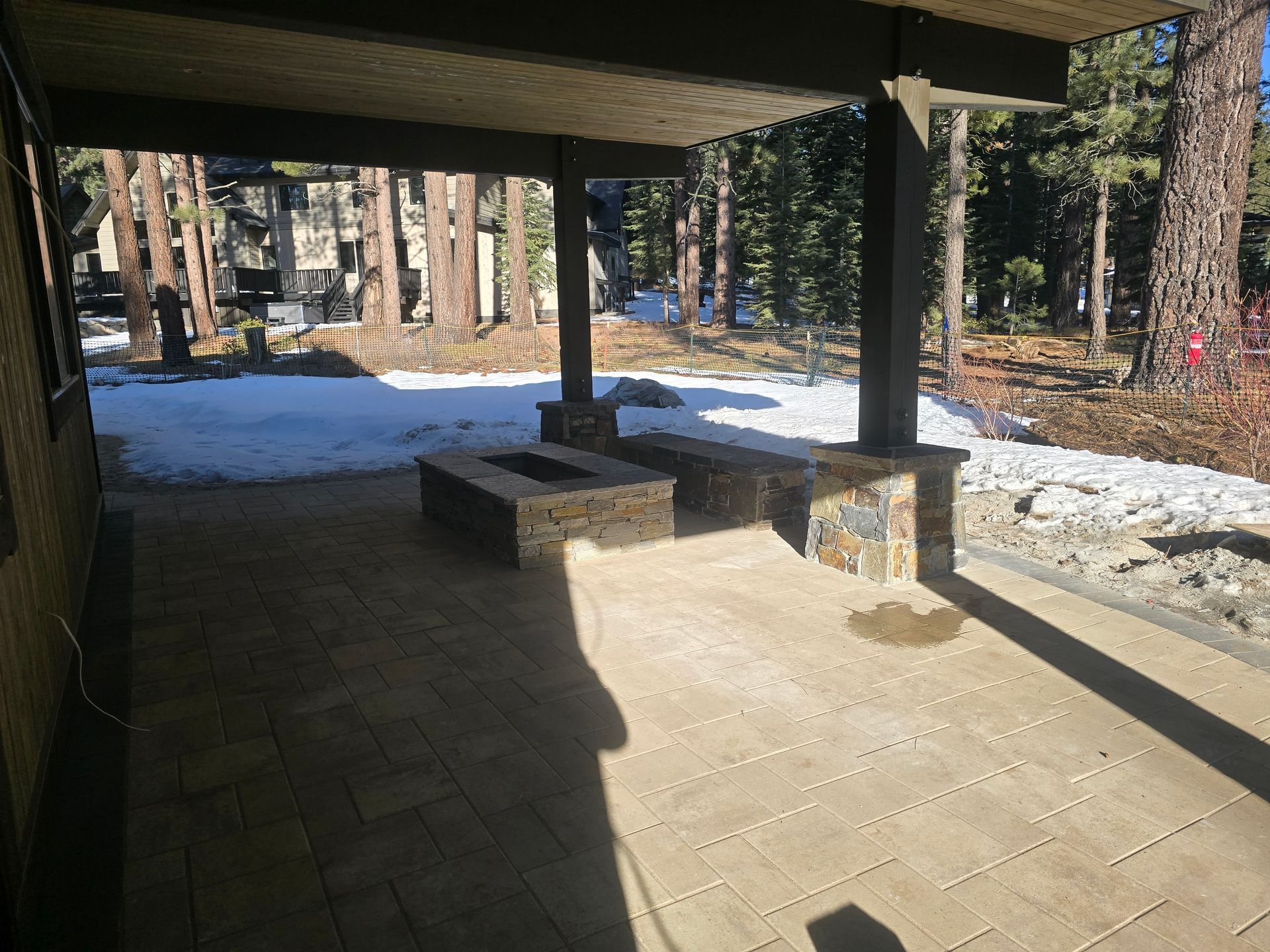 Covered patio with stone pavers, benches, and snow-covered ground beside a wooded area