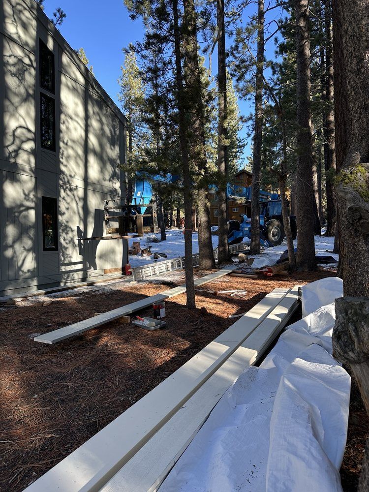Snowy construction site beside a building with tall pine trees and stacked lumber.