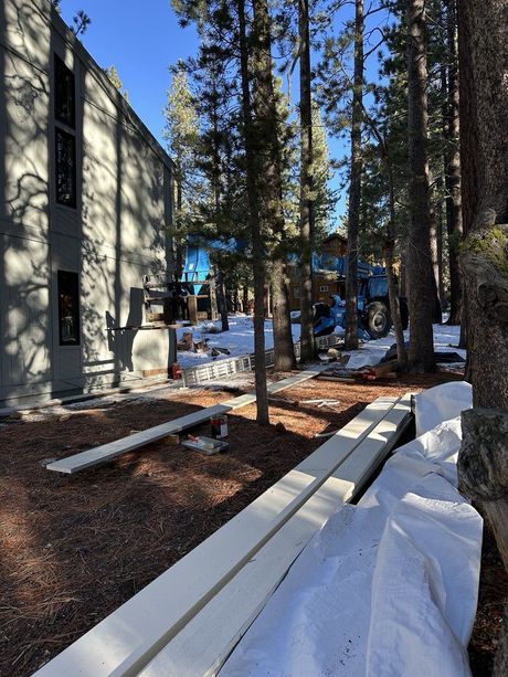 Snowy construction site beside a building with tall pine trees and stacked lumber.