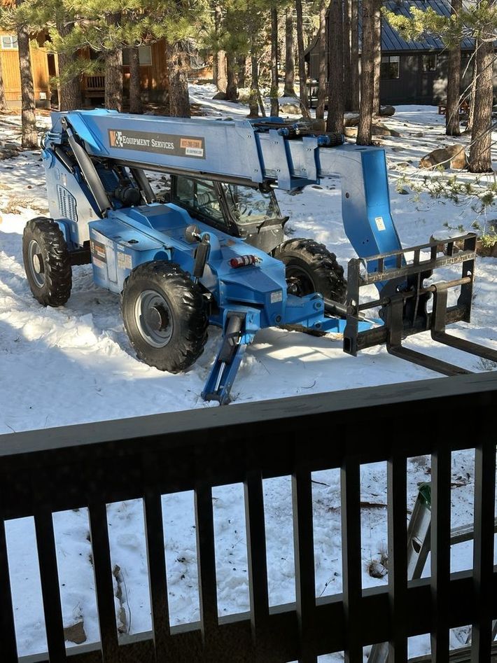 Blue construction lift parked in a snowy yard beside a wooden deck railing.