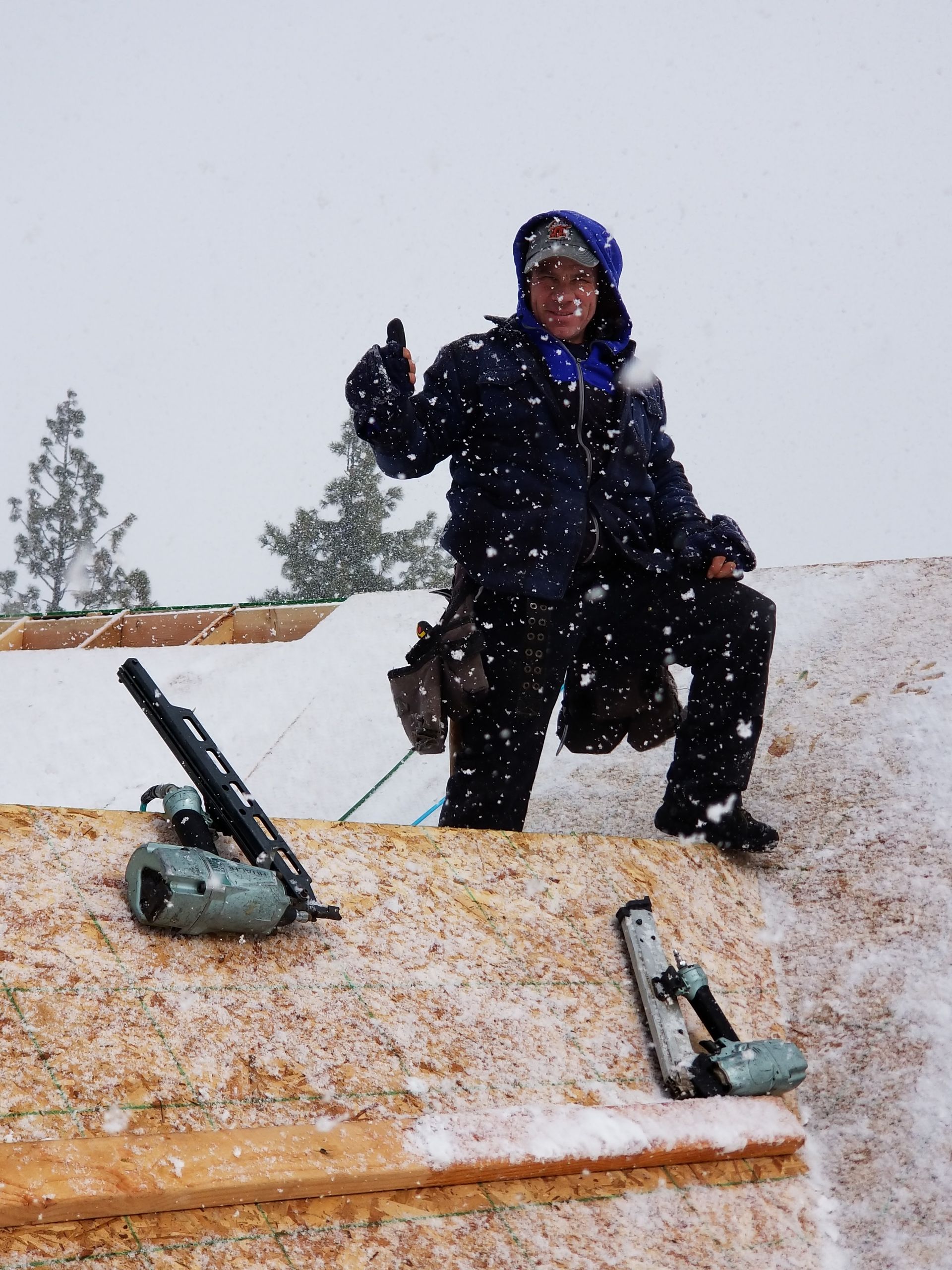 Person kneeling on a snowy rooftop holding a snow tool, with two tools lying nearby.