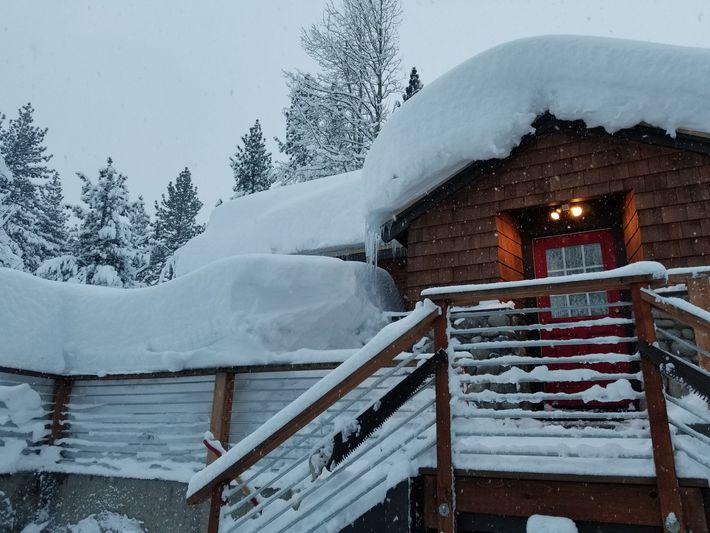 Snow-covered wooden cabin with a lit porch light and stairs in a winter forest