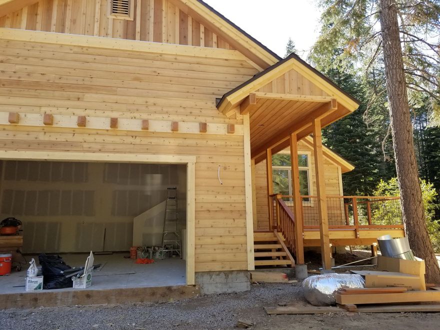 Wood house under construction with open garage and front porch steps, surrounded by trees