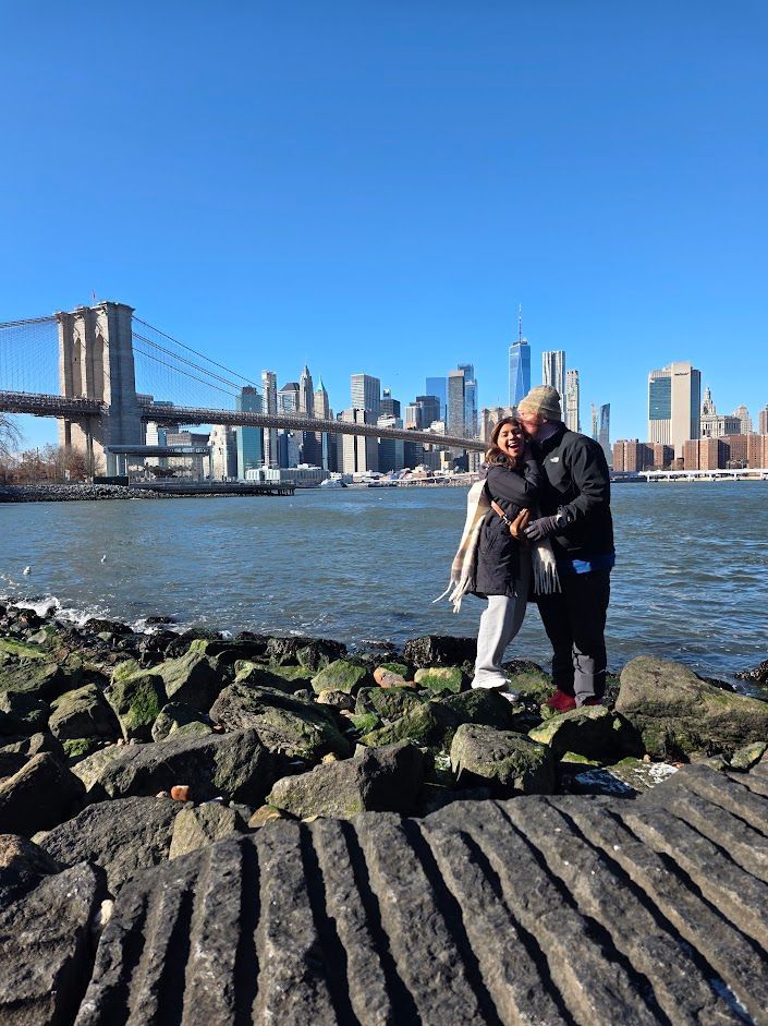 Couple hugging by rocks, Brooklyn Bridge and NYC skyline in background, sunny day.
