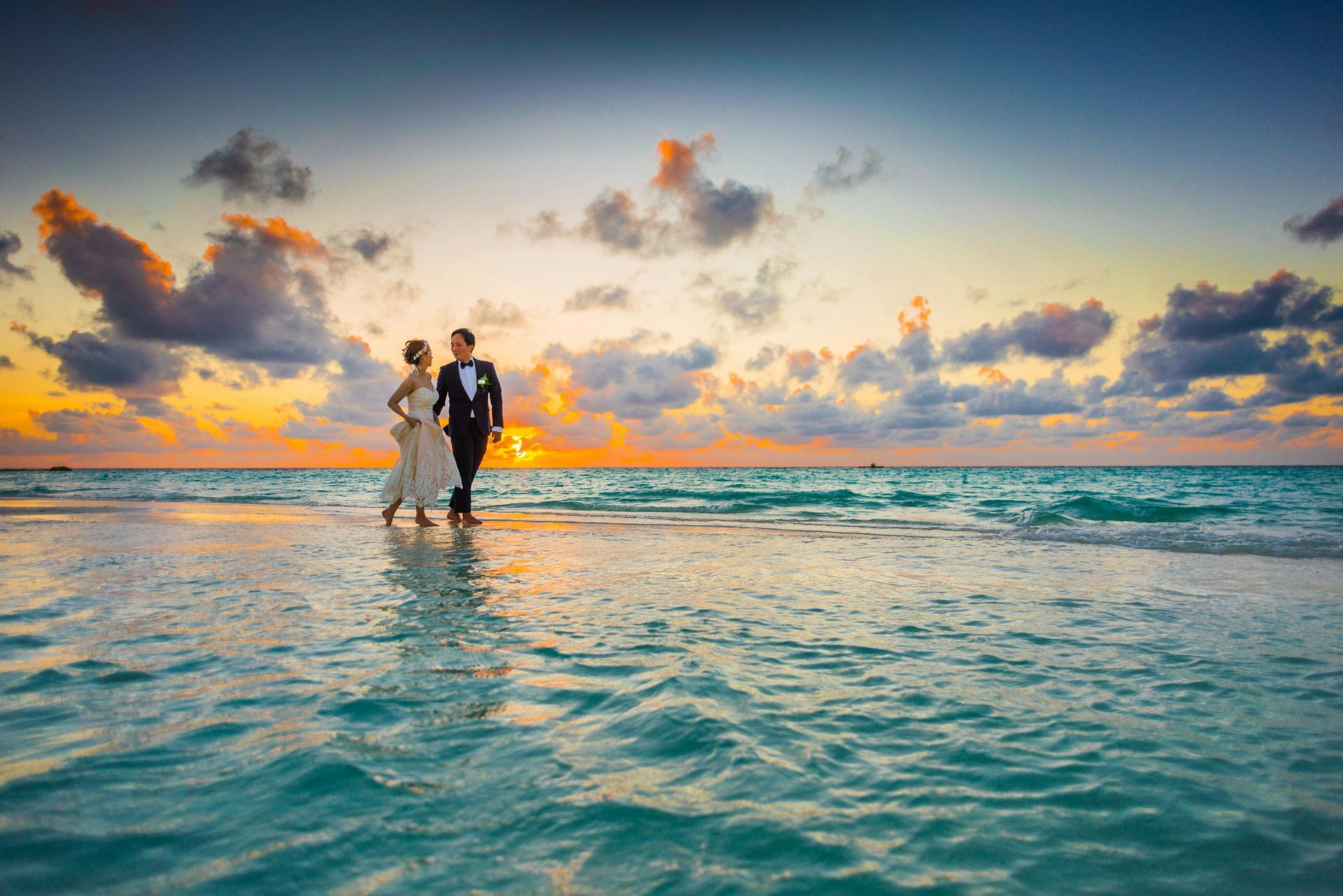 Bride and groom walk on beach at sunset, waves lapping at their feet. Dramatic sky with orange and blue hues.