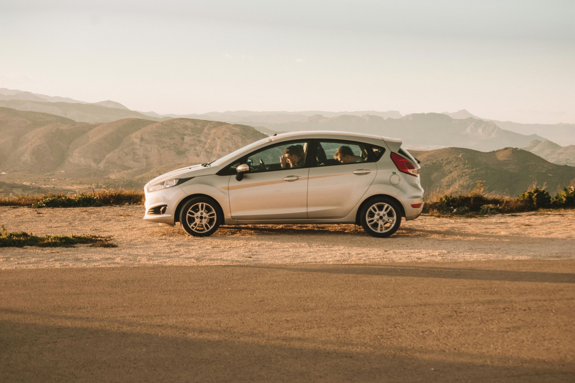 White hatchback car driving on a mountain road at sunset, with passengers inside and hills in the background