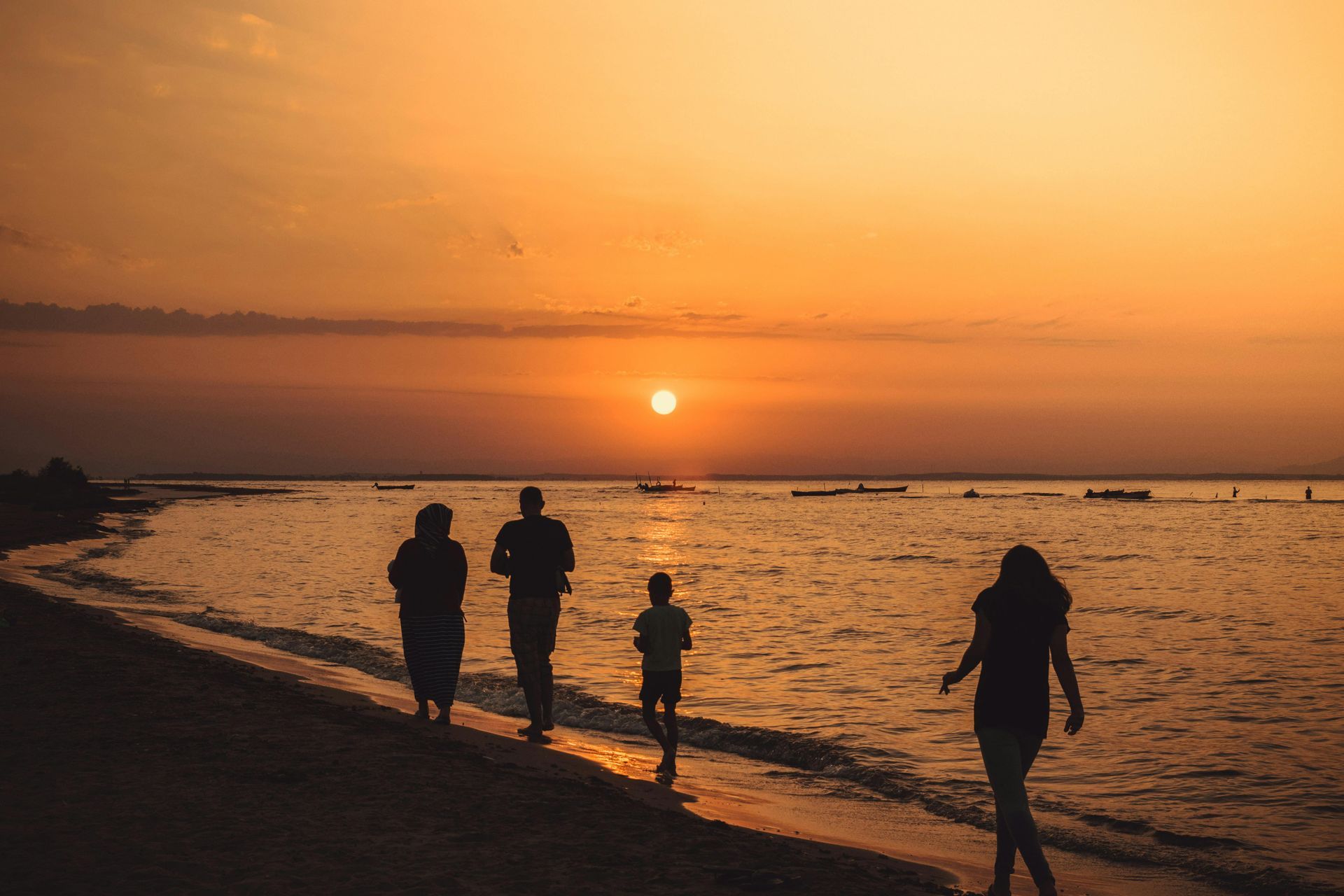 Silhouetted family walking on beach at sunset: orange sky, calm water.