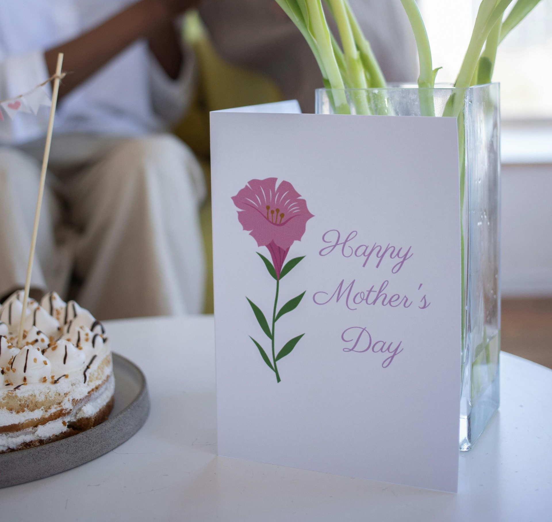 Mother's Day card with pink flower beside a cake on a table, with a vase of flowers in the background