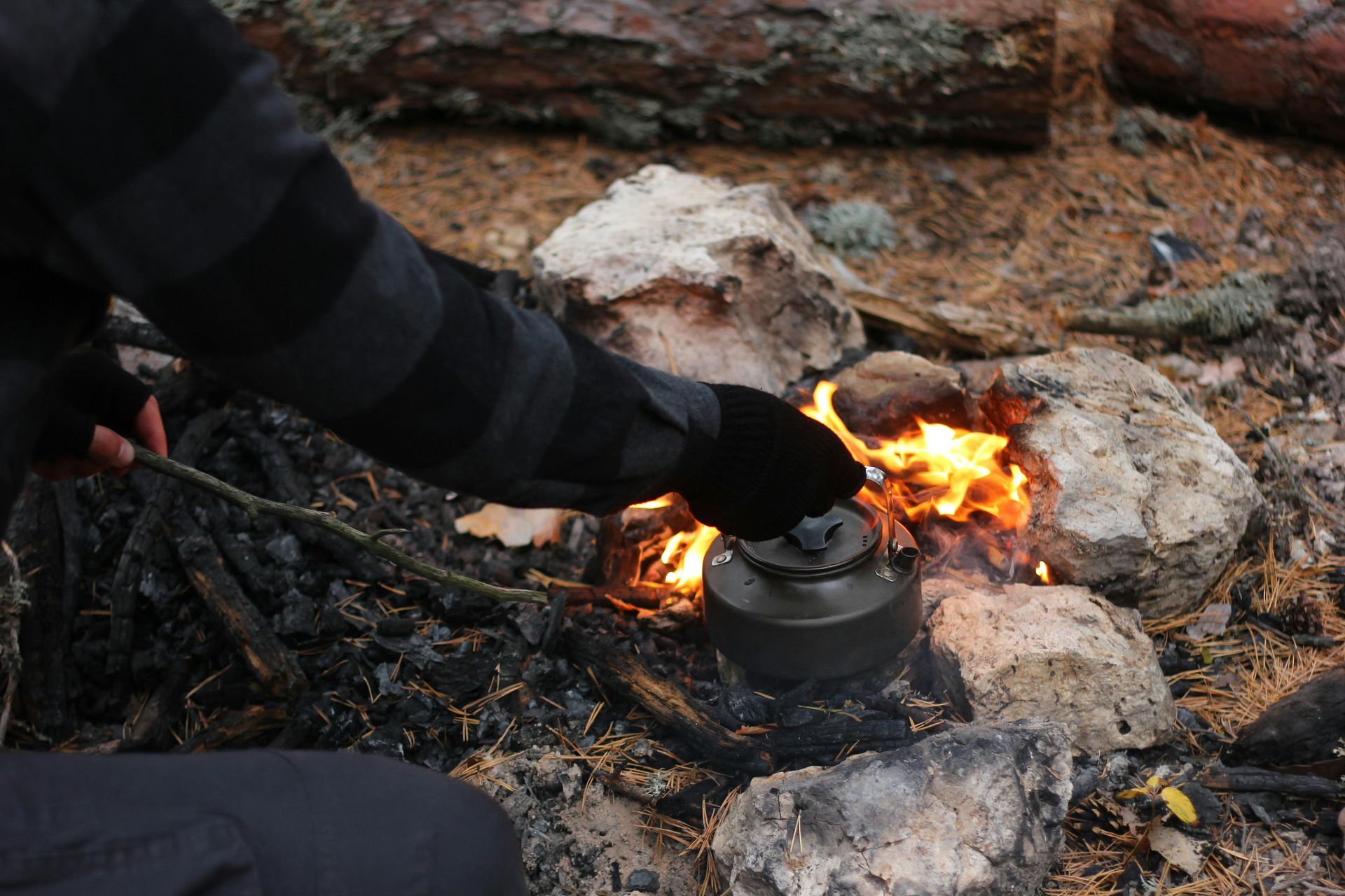 Person tending a campfire, heating a kettle on flames surrounded by rocks and charred wood.