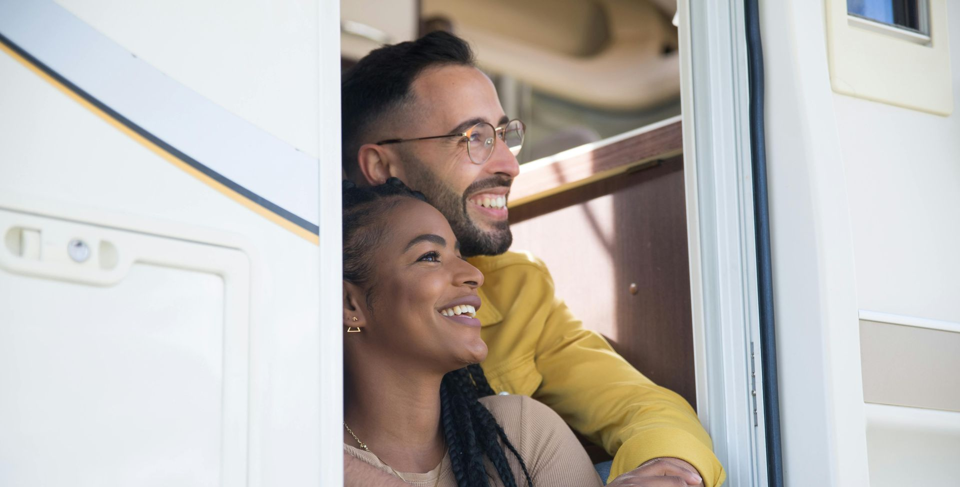 Couple smiling and looking out of a recreational vehicle. Man in yellow jacket, woman with braided hair.