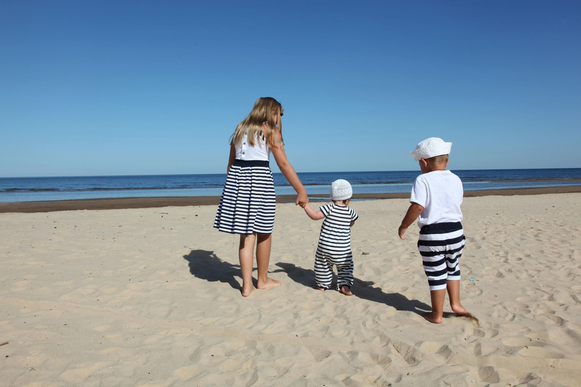 Woman and two children in sailor outfits walking on a sandy beach towards the ocean on a sunny day.