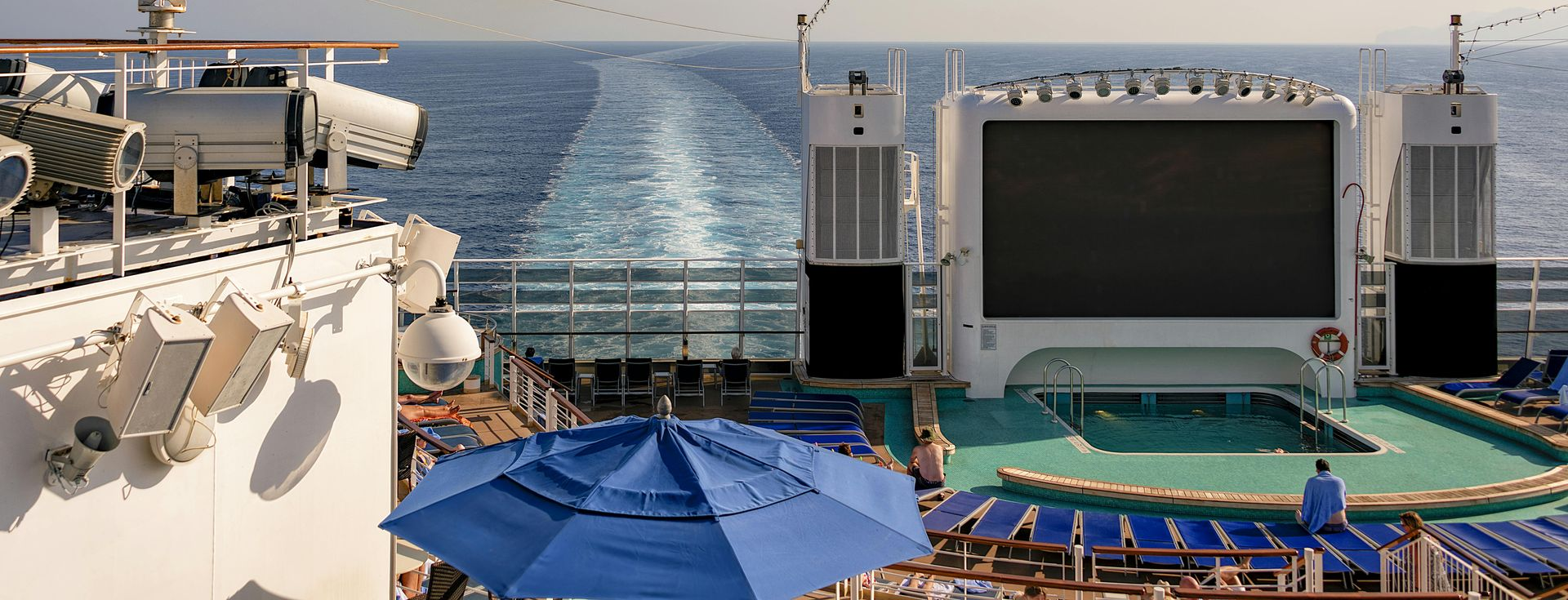 Cruise ship deck with ocean view, large screen, blue umbrella, lounge chairs.