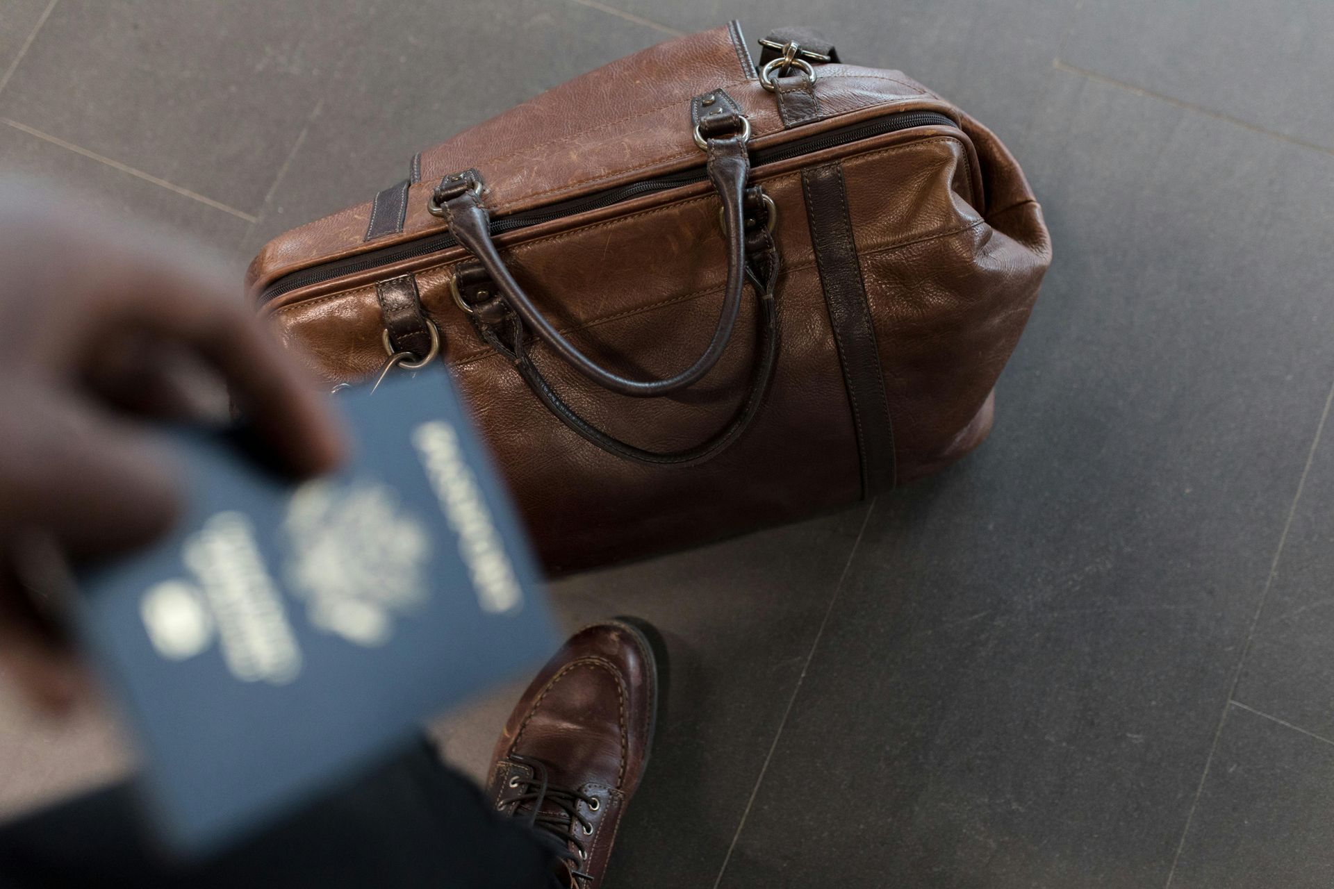 Person holding a blue passport next to a brown leather travel bag on a gray floor.