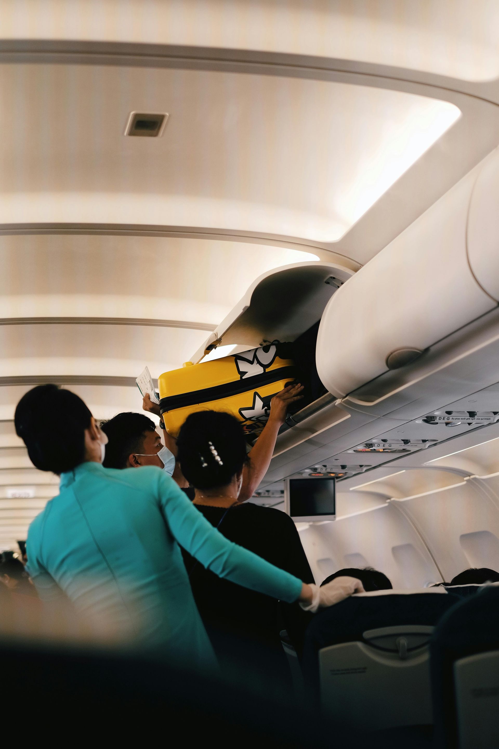Flight attendants assisting a passenger with luggage in the overhead compartment on a plane.
