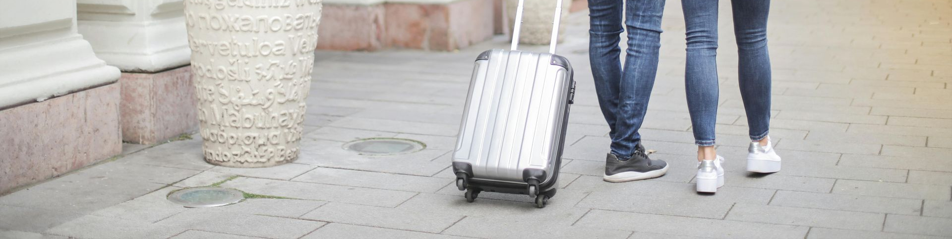 Two people in jeans stand on a paved walkway next to a rolling suitcase.