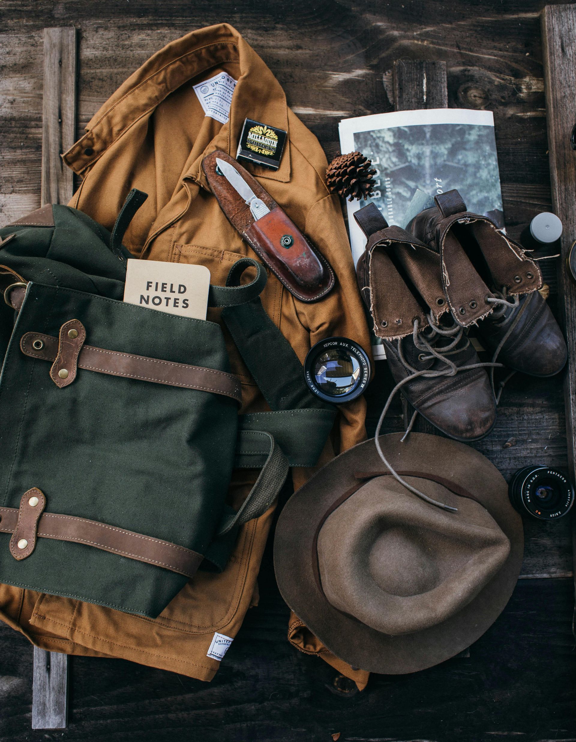 Outdoor gear laid out on wooden planks: backpack, jacket, boots, hat, binoculars, knife. Earth tones.