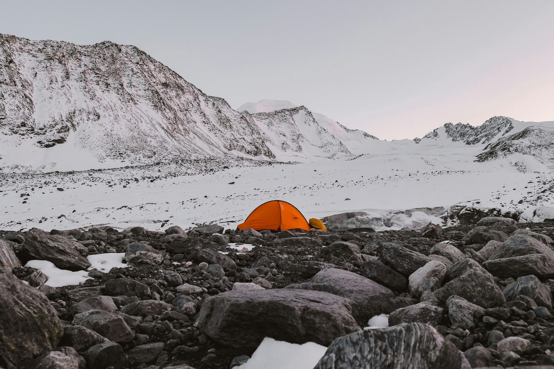 Orange tent on a rocky, snowy landscape, mountains in the background.