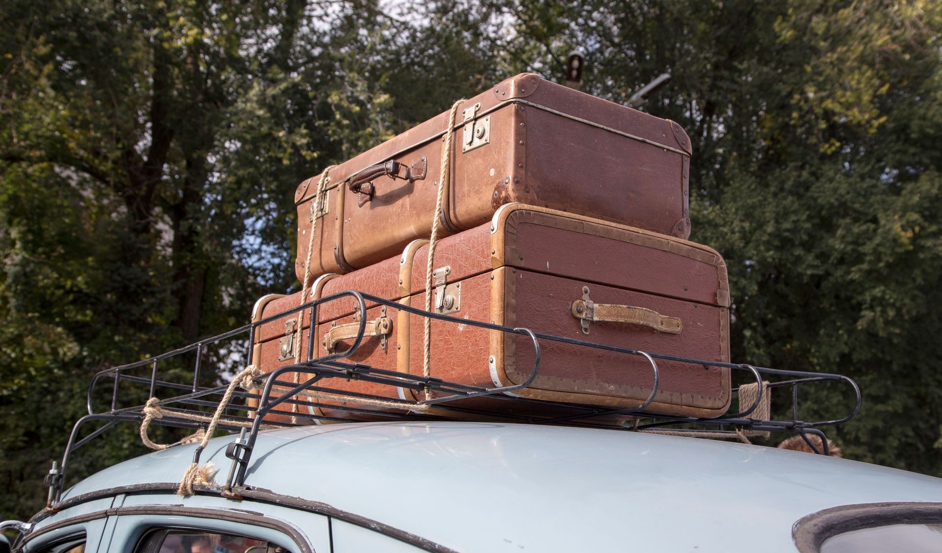 Two brown suitcases strapped to a car roof rack outdoors under trees.