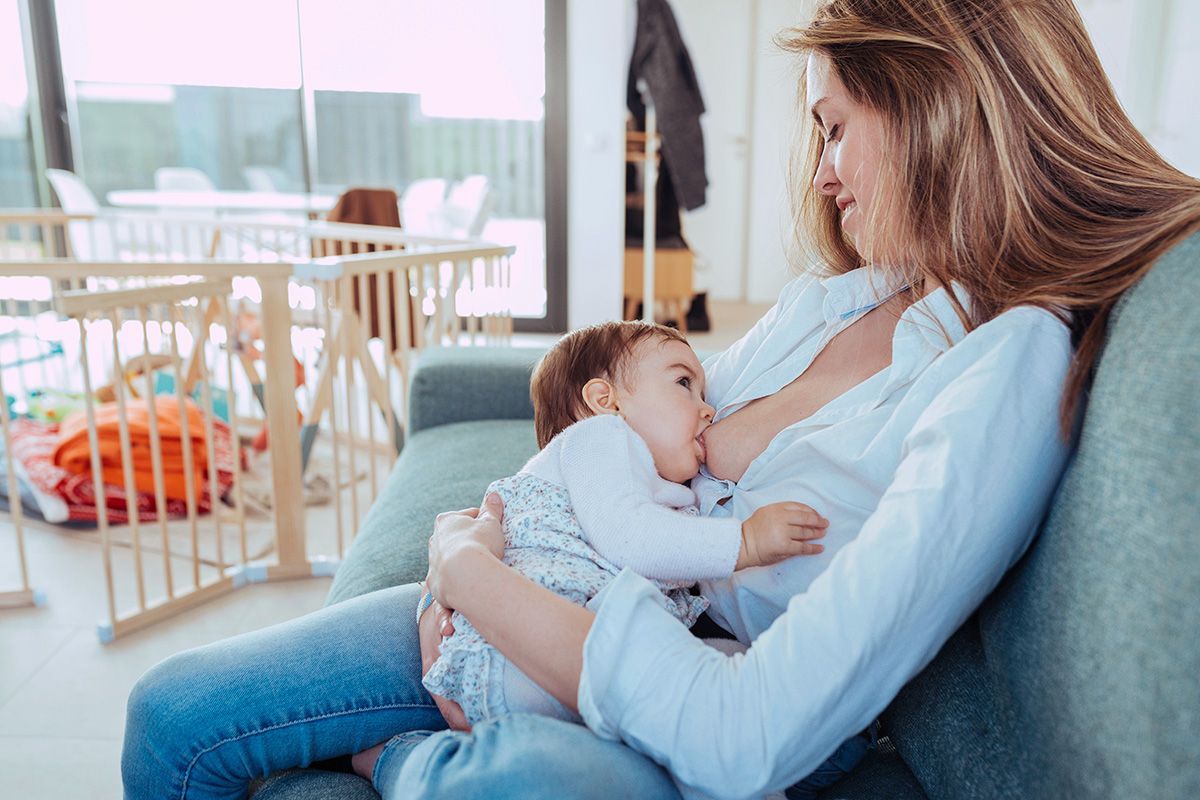 Woman breastfeeding a baby on a couch, indoors. A wooden playpen is visible.