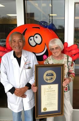 Two people in white coats and a woman holding a plaque stand beside a large orange mascot.