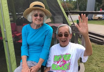 Two smiling people sitting outdoors, one waving, in colorful casual clothes and a sun hat